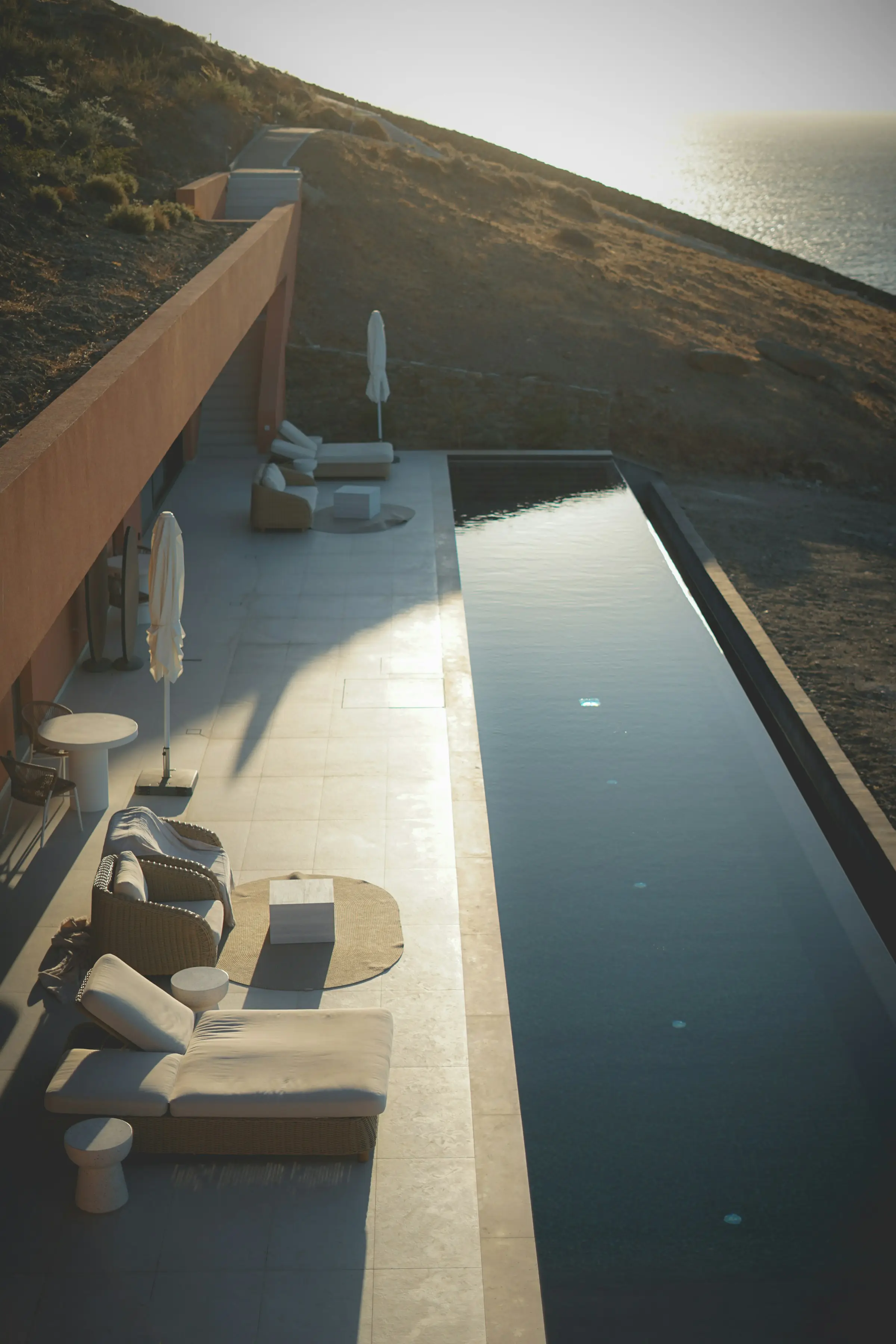 Modern outdoor patio with cushioned lounge chairs and umbrellas beside a narrow infinity pool overlooking a hillside and ocean at sunset.