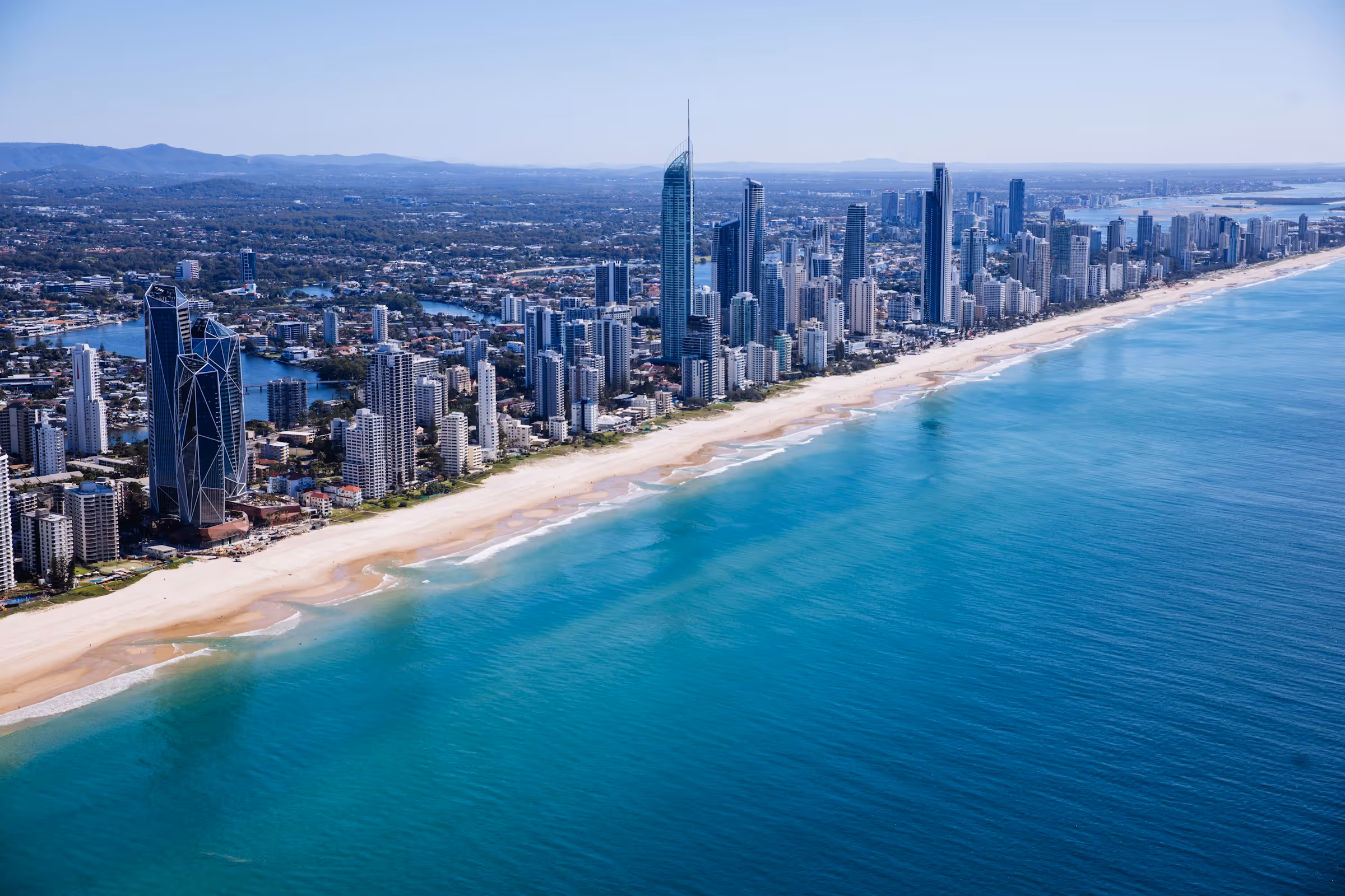 Aerial view of a coastal city skyline with tall skyscrapers along a sandy beach and turquoise ocean.