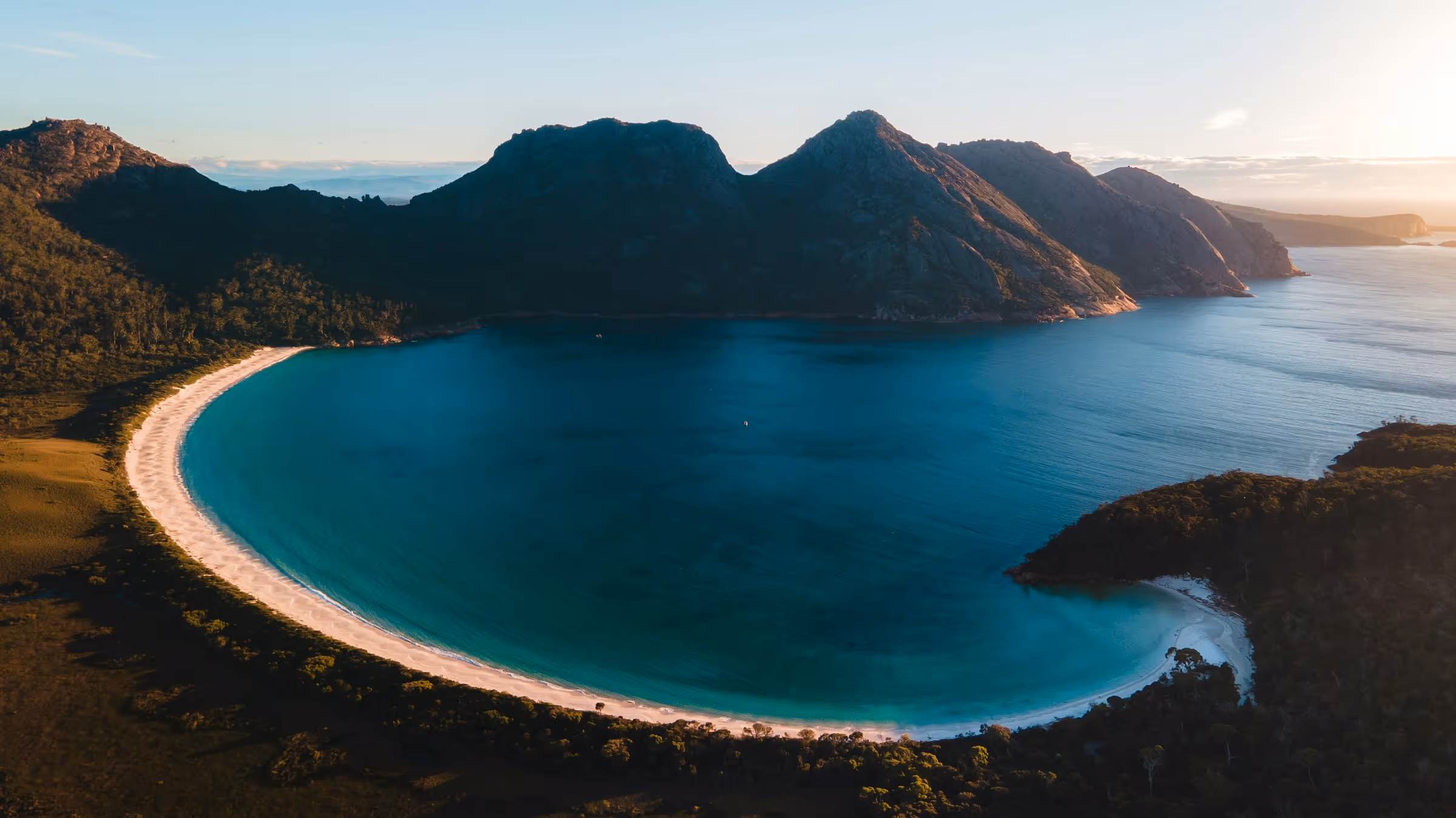 A curved sandy beach with clear blue water surrounded by forested hills under a bright sky at sunset.