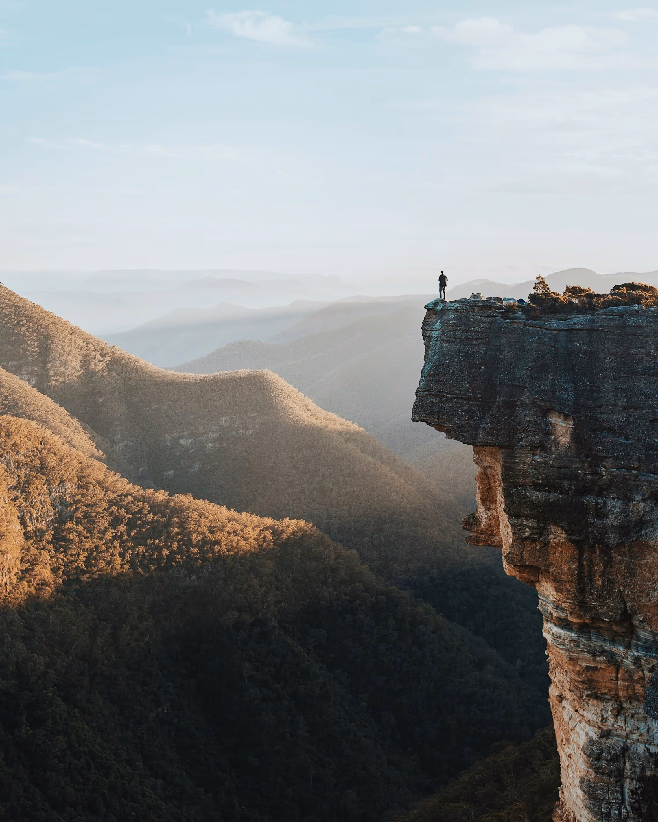 Person standing on the edge of a high rocky cliff overlooking a vast mountainous landscape at sunrise or sunset.