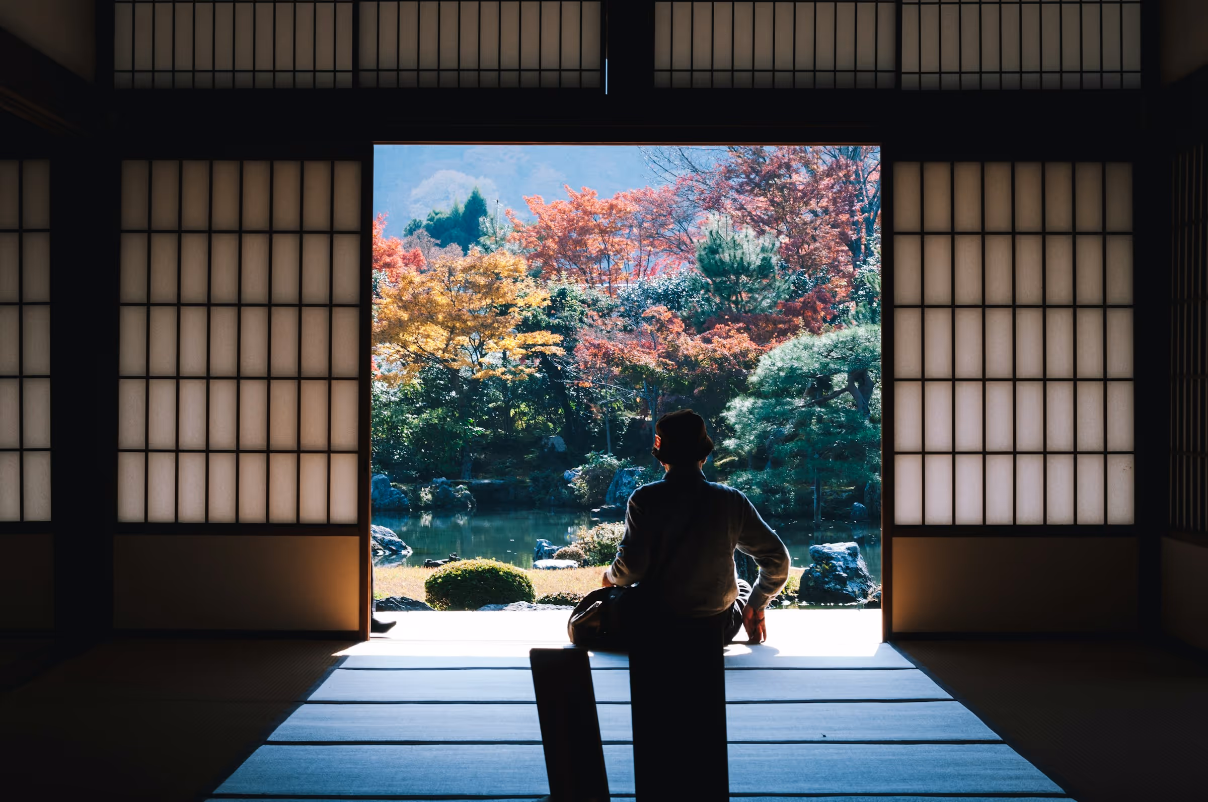 Person sitting inside a traditional Japanese-style room facing an open doorway with a view of a colorful autumn garden and pond.