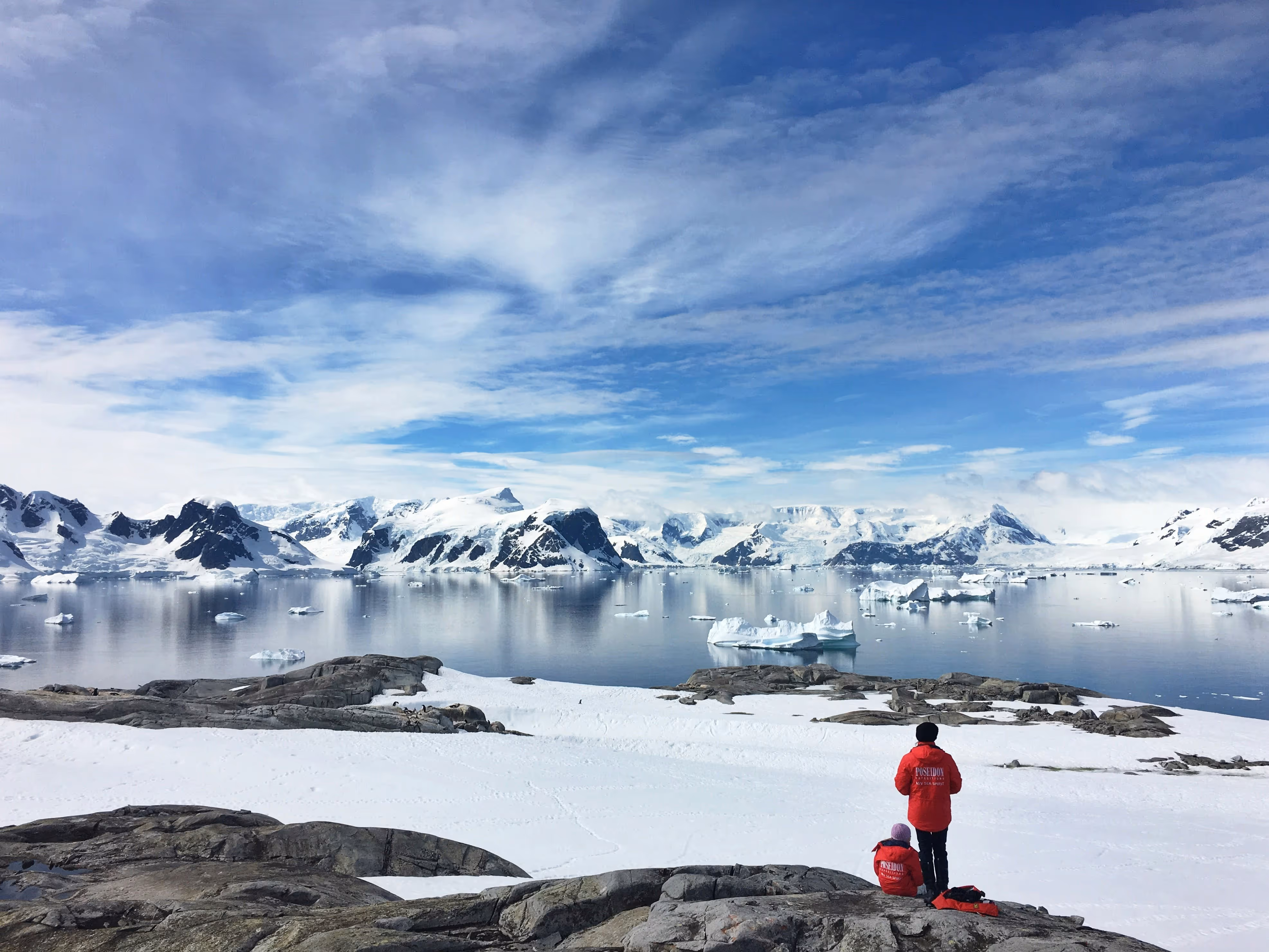 Two people wearing red jackets overlooking icy mountains and floating icebergs on a calm body of water under partly cloudy blue sky.