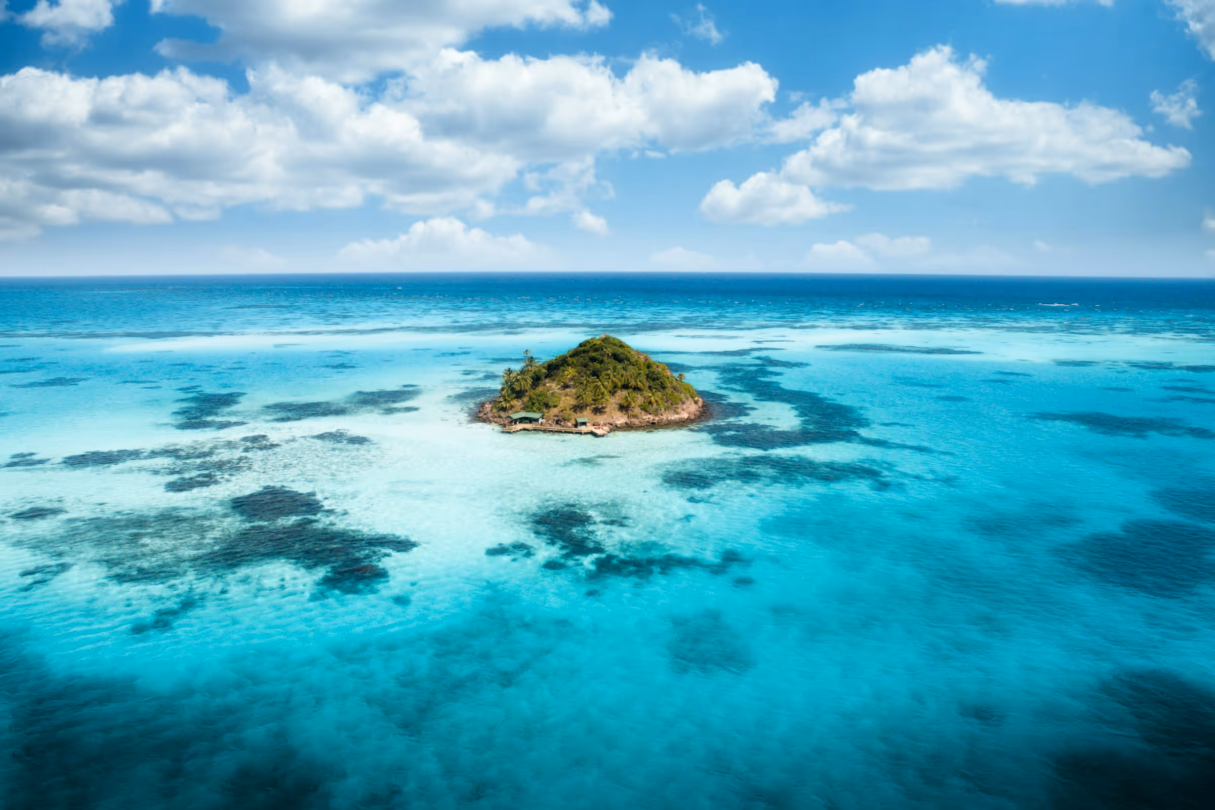 Small lush green island surrounded by clear turquoise ocean under a partly cloudy blue sky.