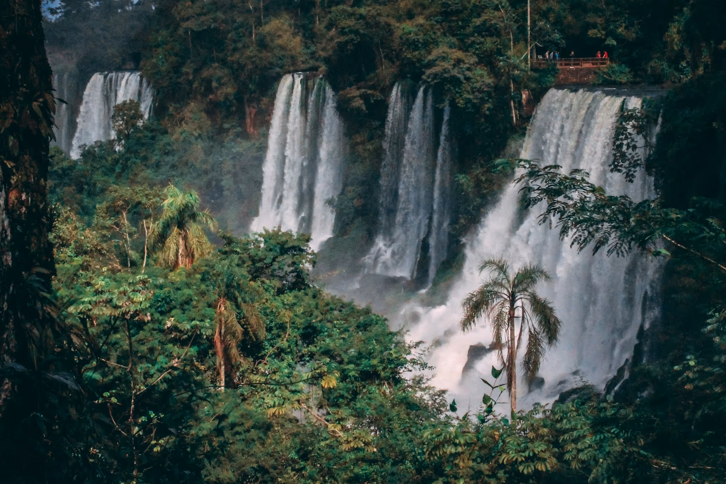Multiple large waterfalls cascading down cliffs surrounded by dense green tropical forest with a viewing platform on the top right.