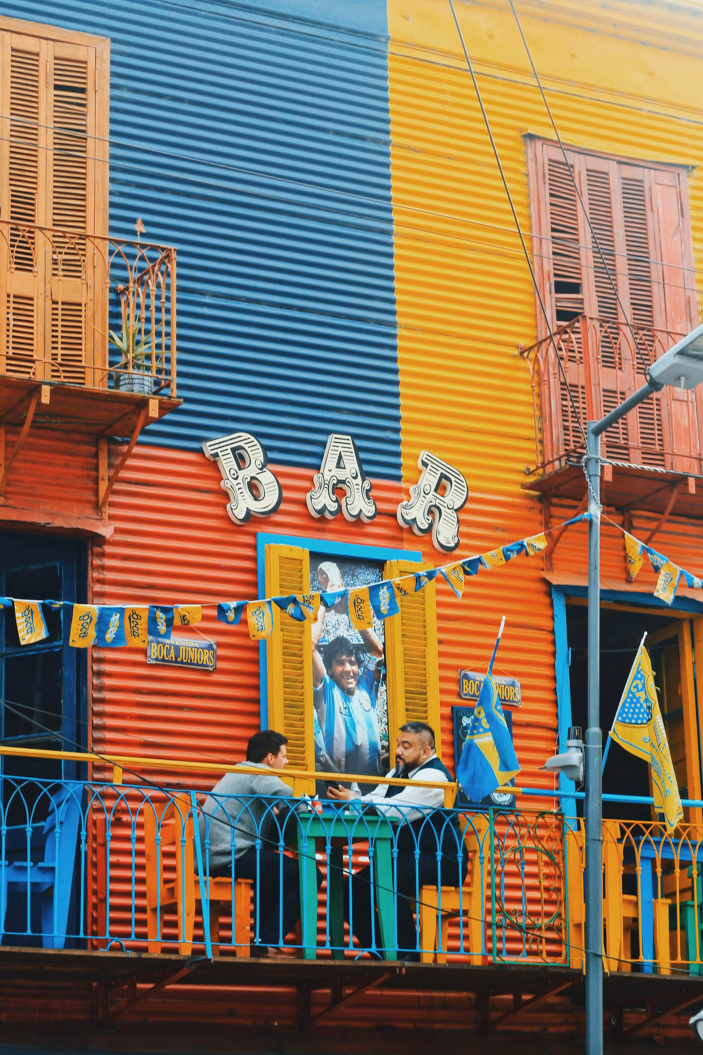 Colorful bar facade painted in blue, red, and yellow with two men sitting at a table on the balcony under a sign that reads 'BAR' and a poster of a soccer player holding a trophy.