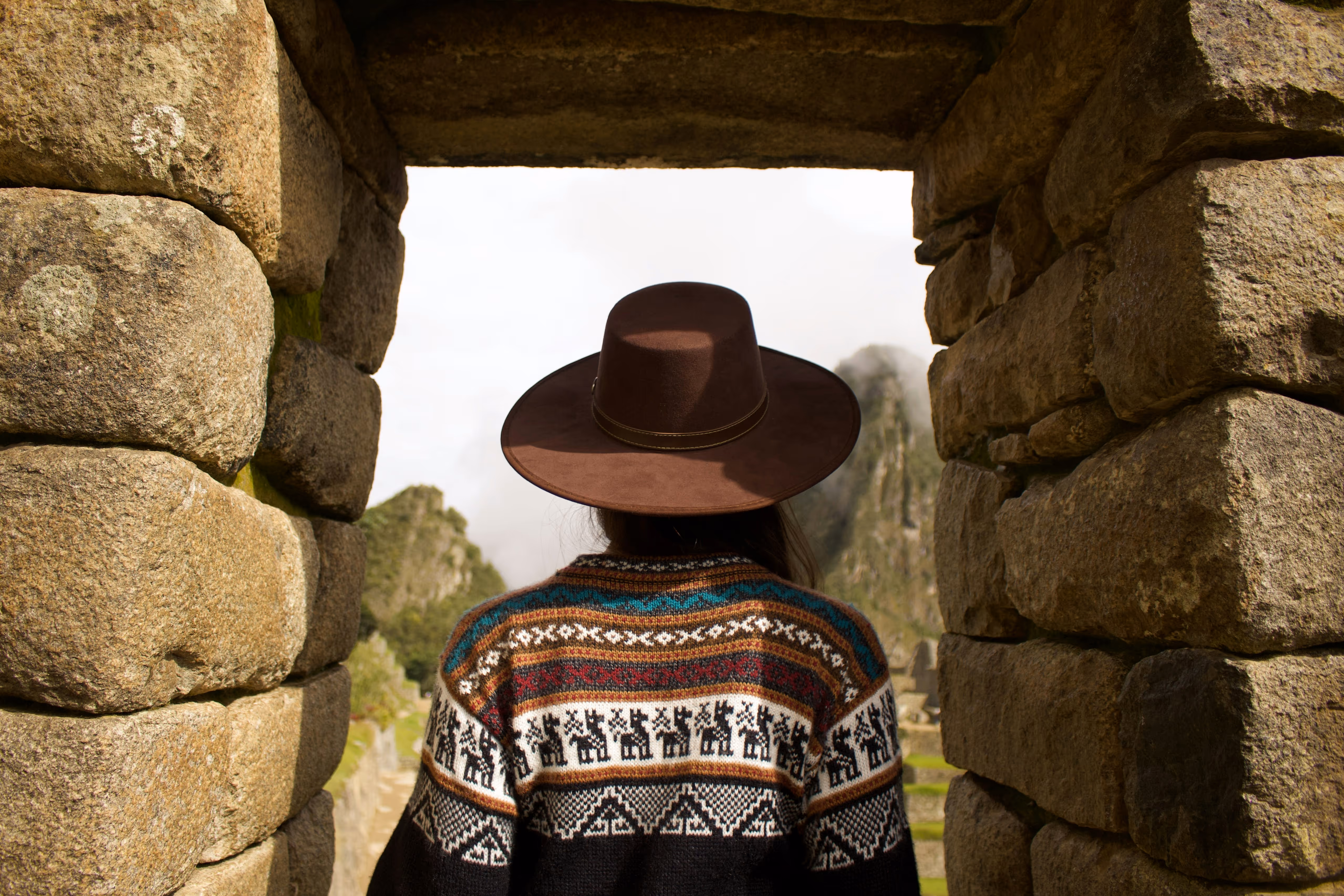 Person wearing a brown hat and patterned sweater looking out through an ancient stone doorway at a mountainous landscape.