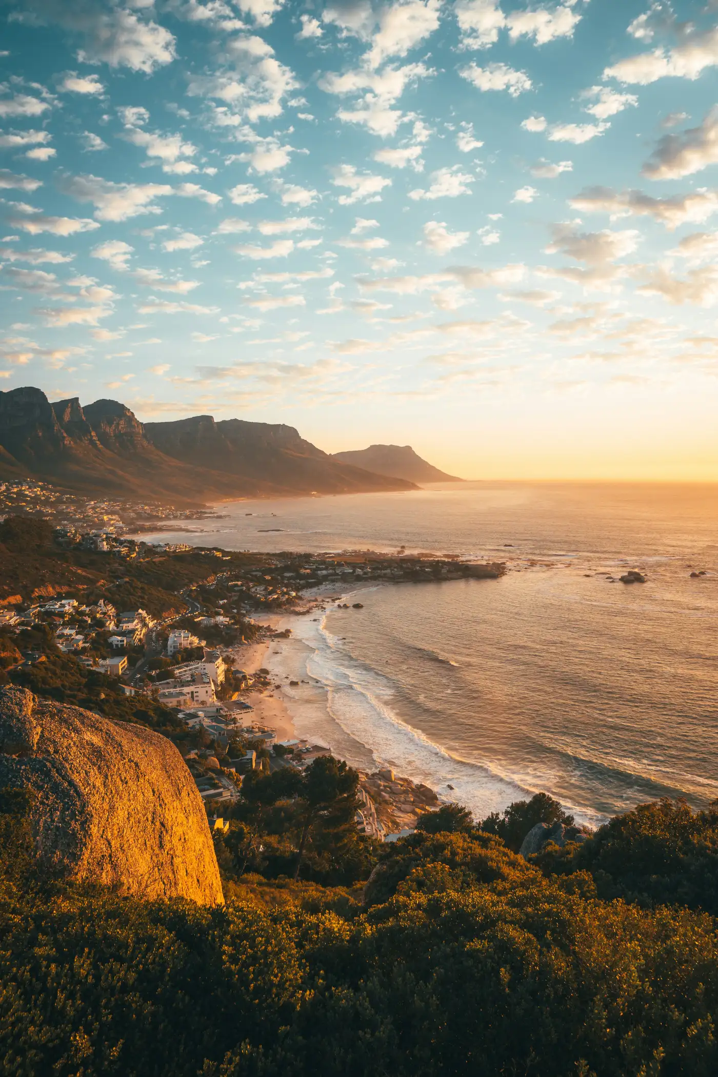 View of a coastal town with sandy beaches, ocean waves, and mountains under a partly cloudy sky at sunset.