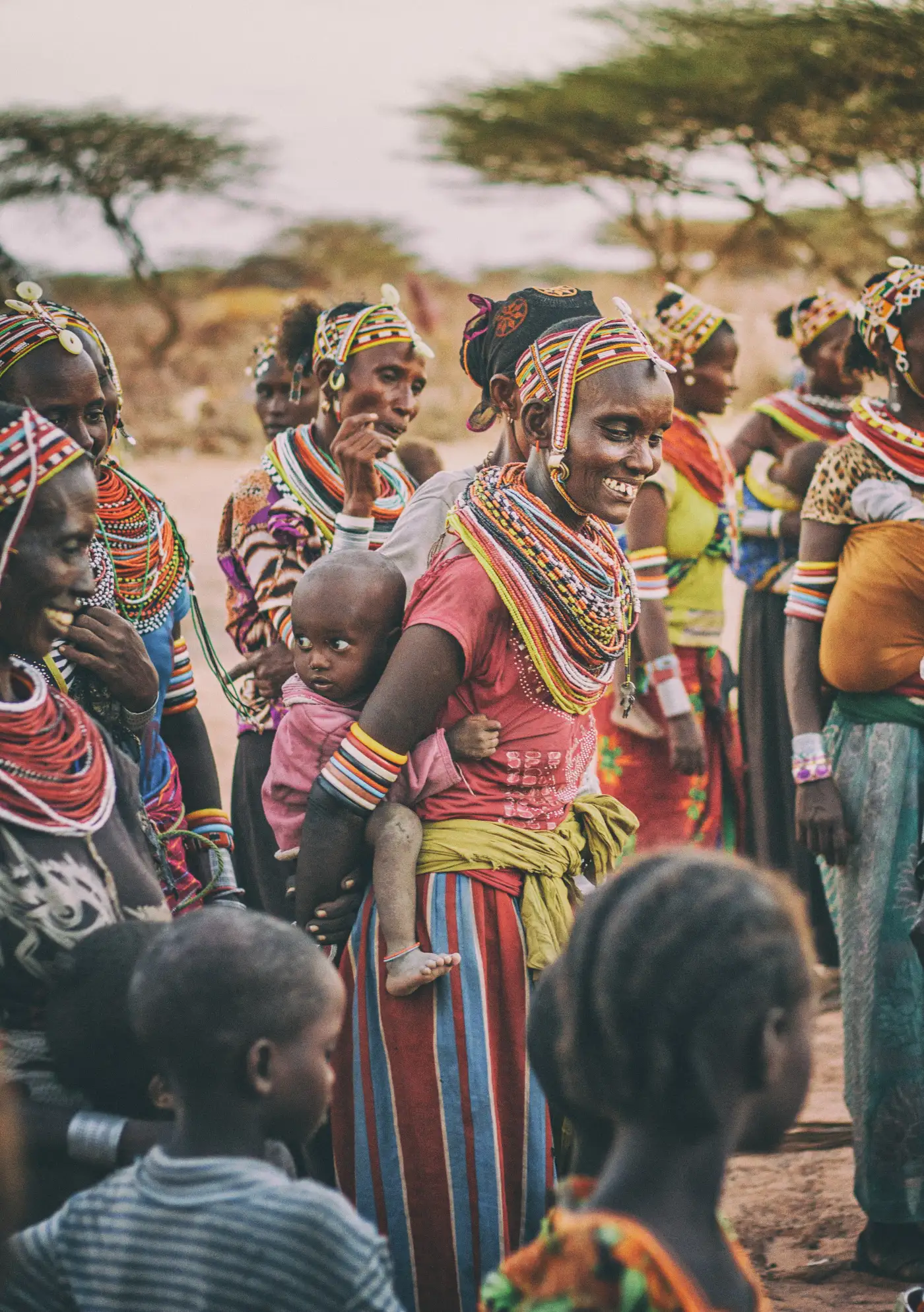 Group of indigenous women dressed in colorful traditional beadwork and clothing, one carrying a child on her back, with children in the foreground.