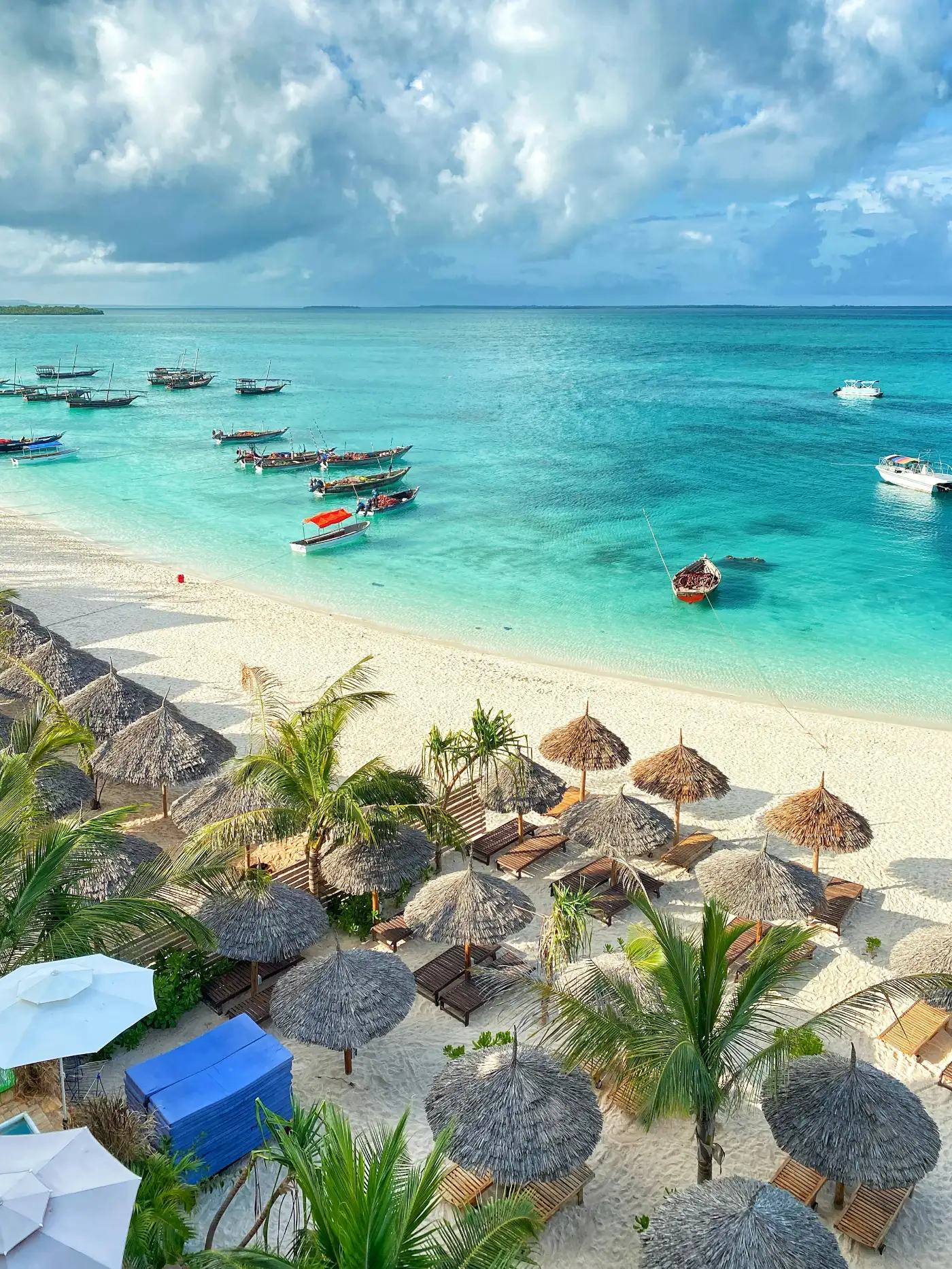 A tropical beach with white sand, several palm trees, thatched umbrellas, lounge chairs, and boats floating on clear turquoise water under a partly cloudy sky.