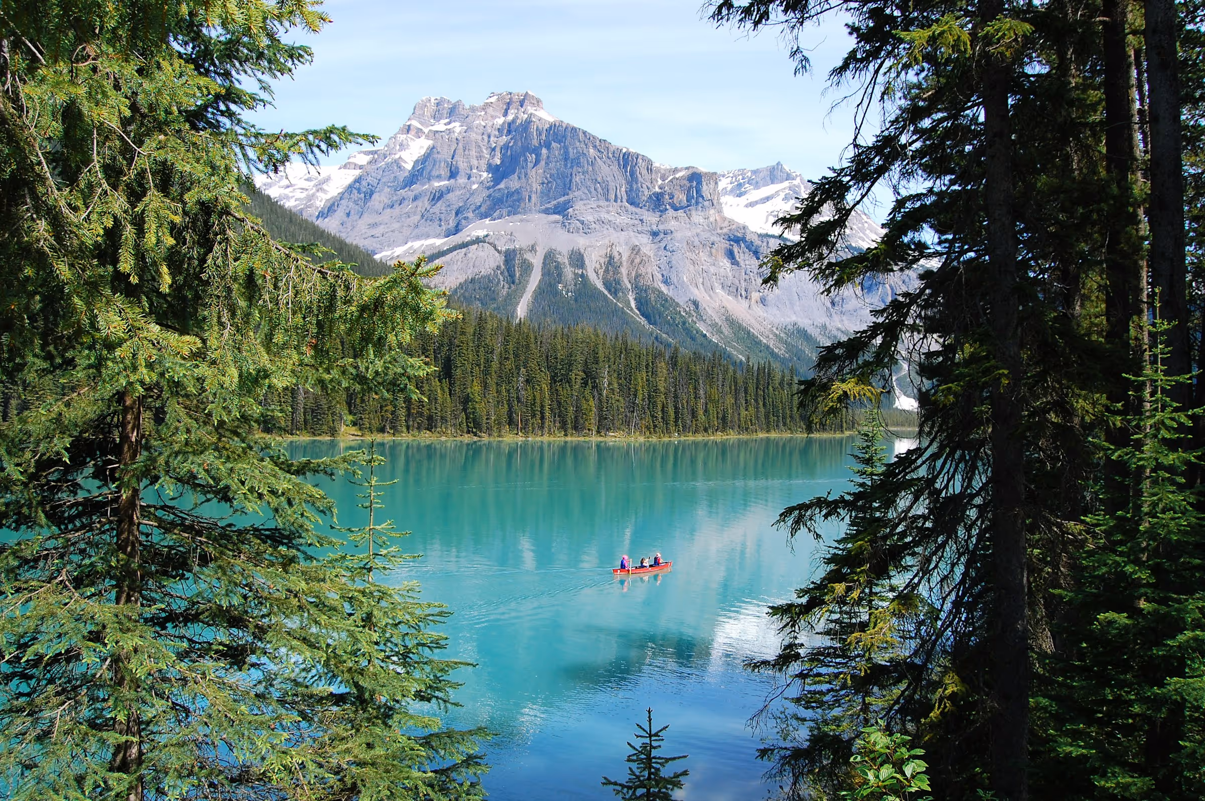 Three people in a red canoe paddling on a turquoise lake surrounded by pine trees with snow-capped mountains in the background.