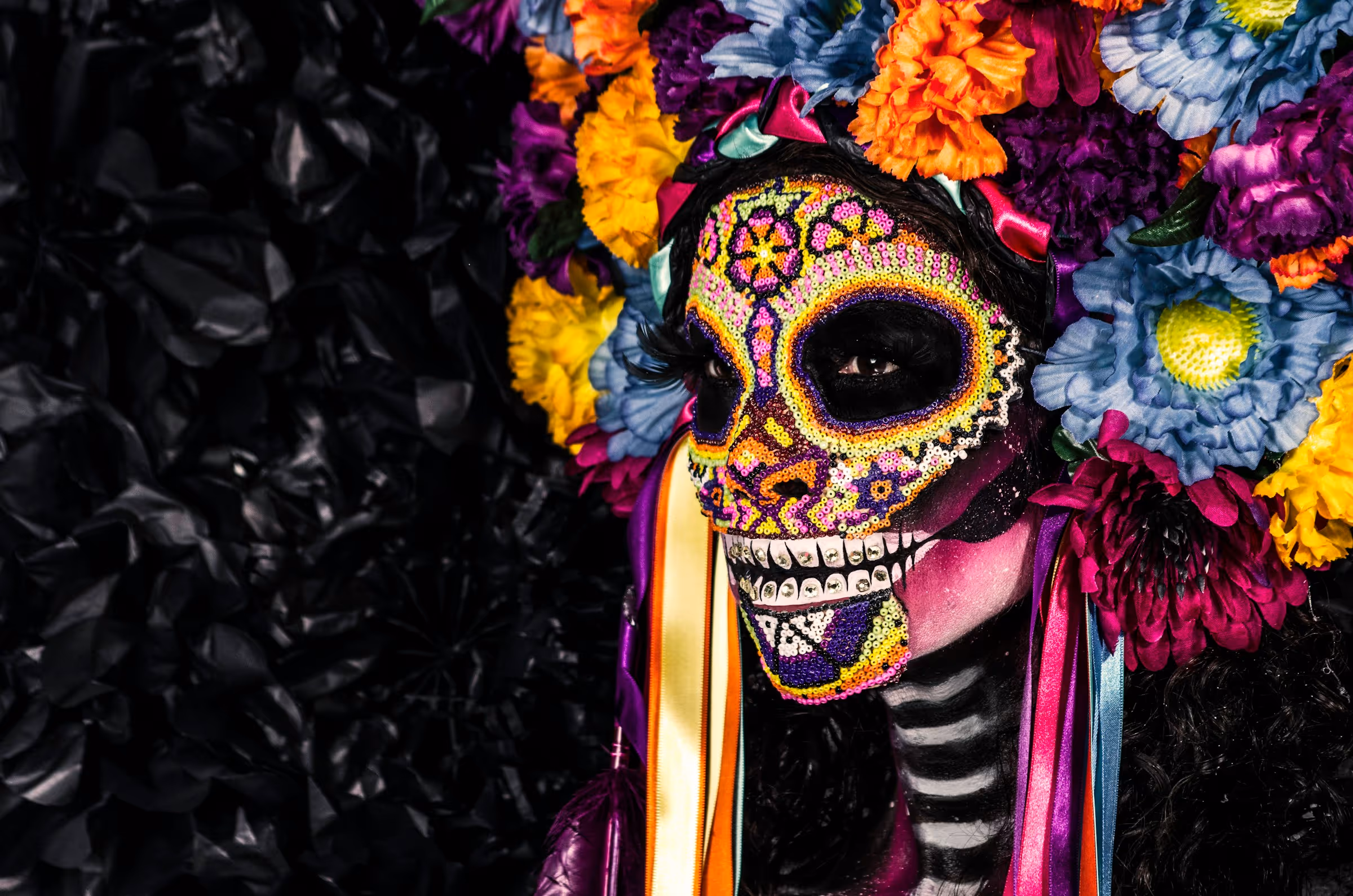Person with colorful Day of the Dead sugar skull face paint and vibrant flower crown against a dark background.
