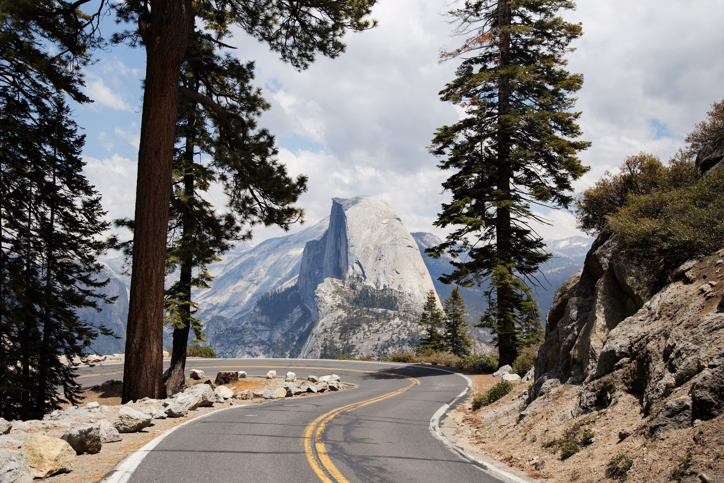 Curving mountain road framed by pine trees with Half Dome rock formation in the background under a cloudy sky.