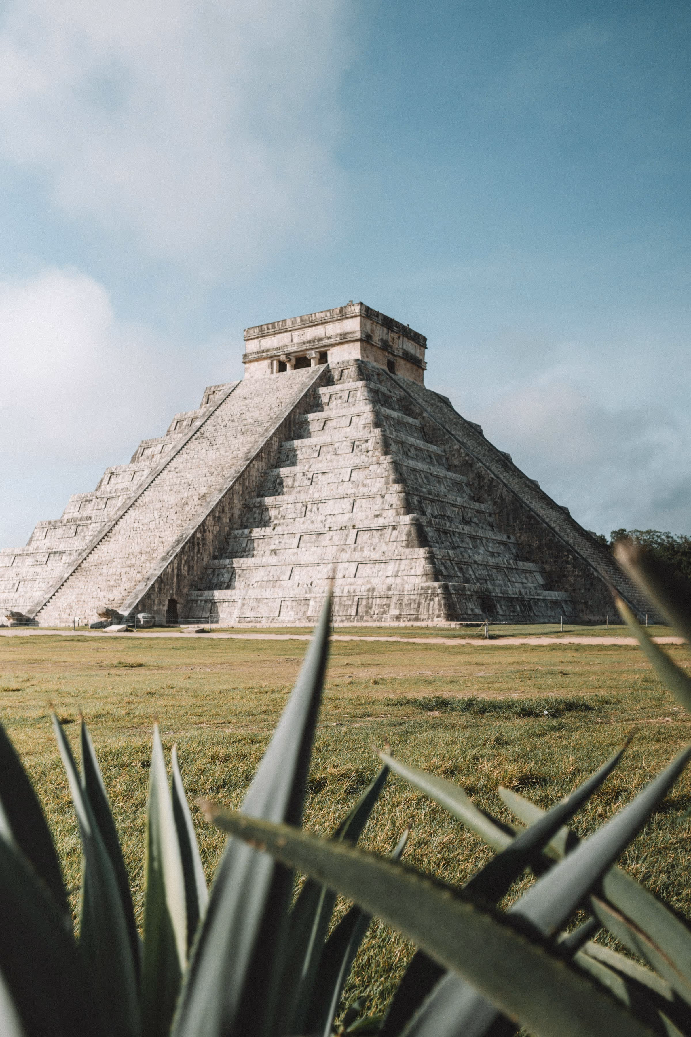 The stone pyramid of Chichen Itza in Mexico with agave plants in the foreground under a partly cloudy sky.
