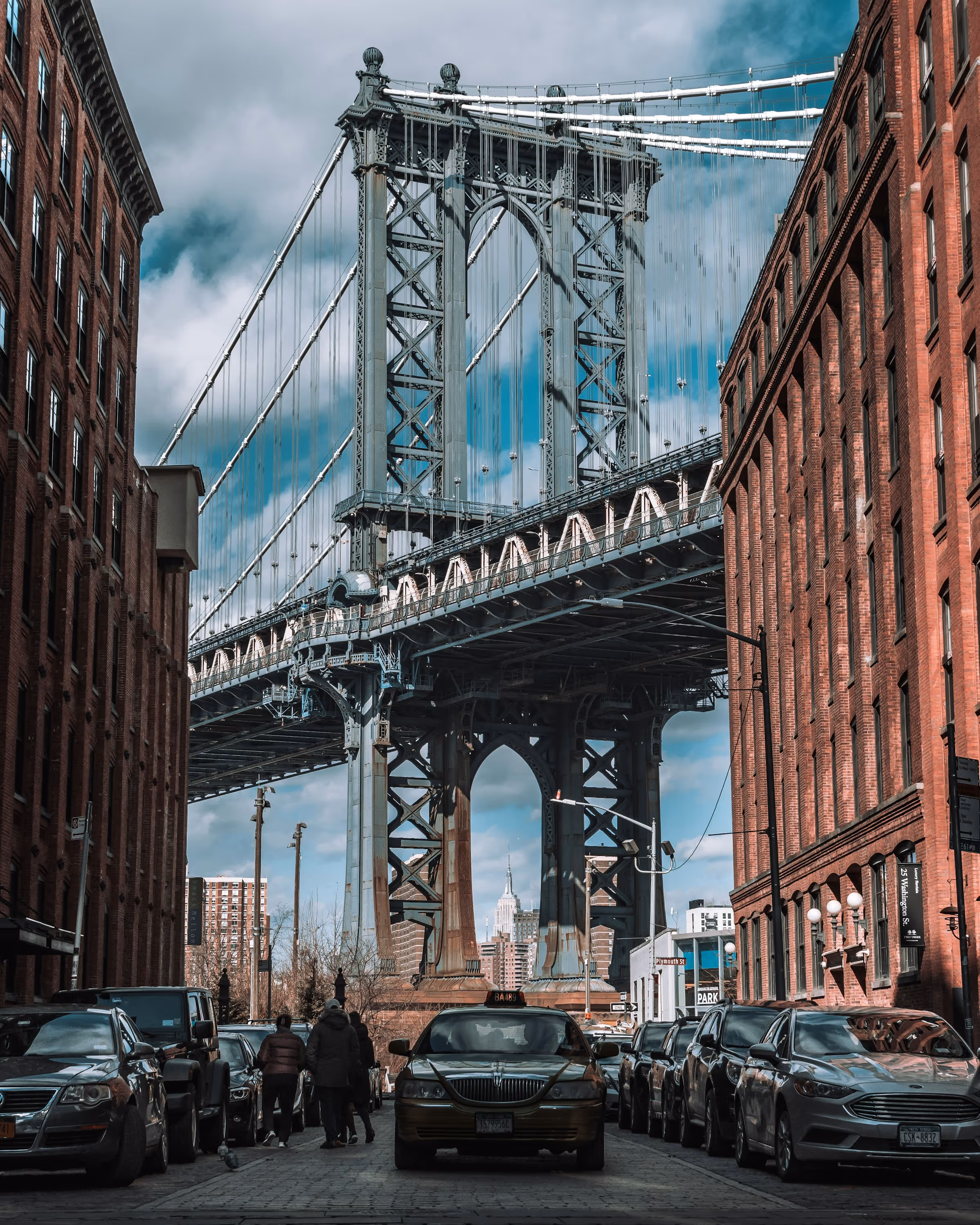 View of the Manhattan Bridge framed by red brick buildings with cars parked on both sides and people walking on the street, under a partly cloudy sky.