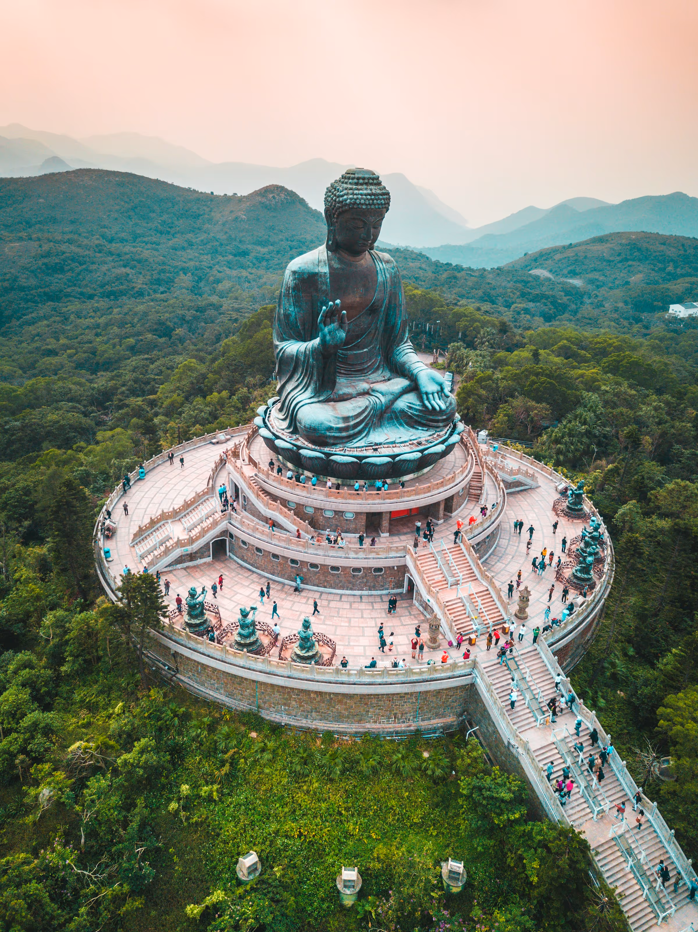 Aerial view of the large bronze Tian Tan Buddha statue on a circular platform surrounded by people and green mountains during sunset.