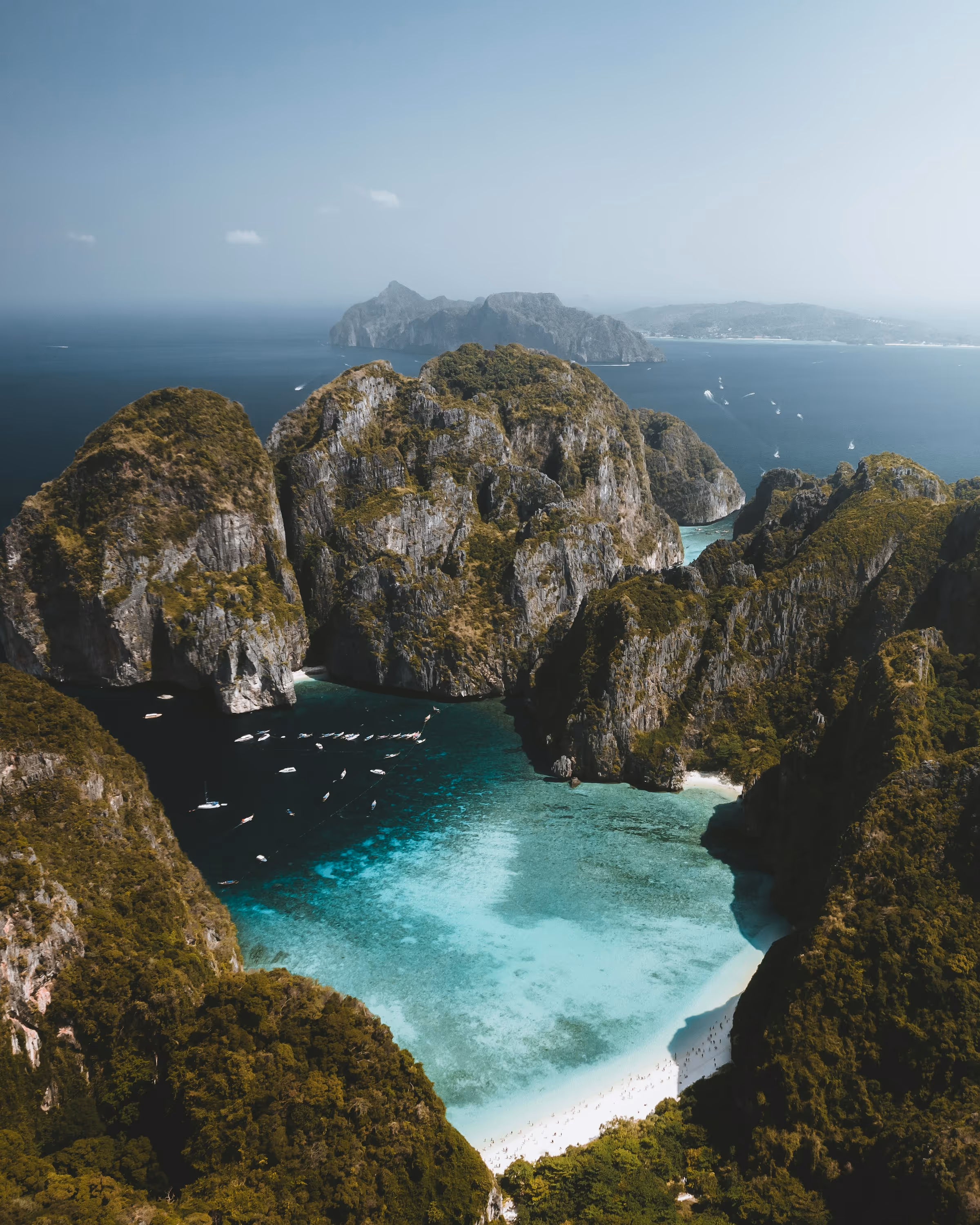 Aerial view of a tropical bay with turquoise water, white sandy beach, and surrounding steep green cliffs with boats anchored near the shore.