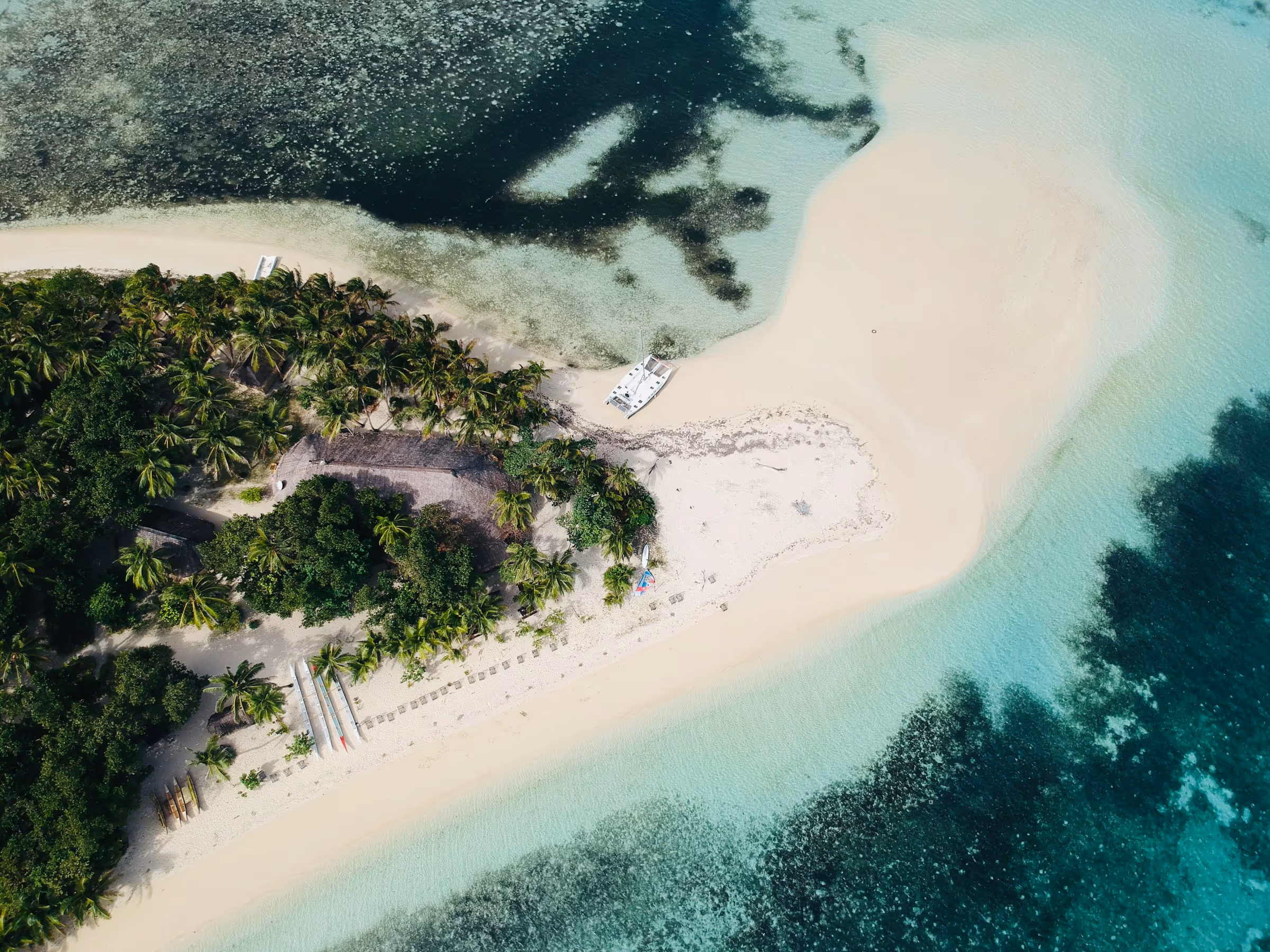 Aerial view of a tropical island with sandy beaches, turquoise water, palm trees, and a docked white boat.