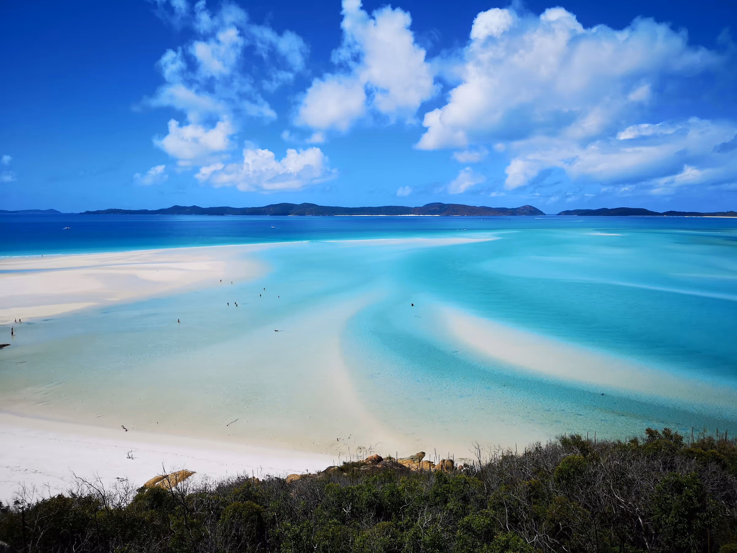 Panoramic view of a tropical beach with white sand bars and turquoise water under a bright blue sky with scattered clouds.