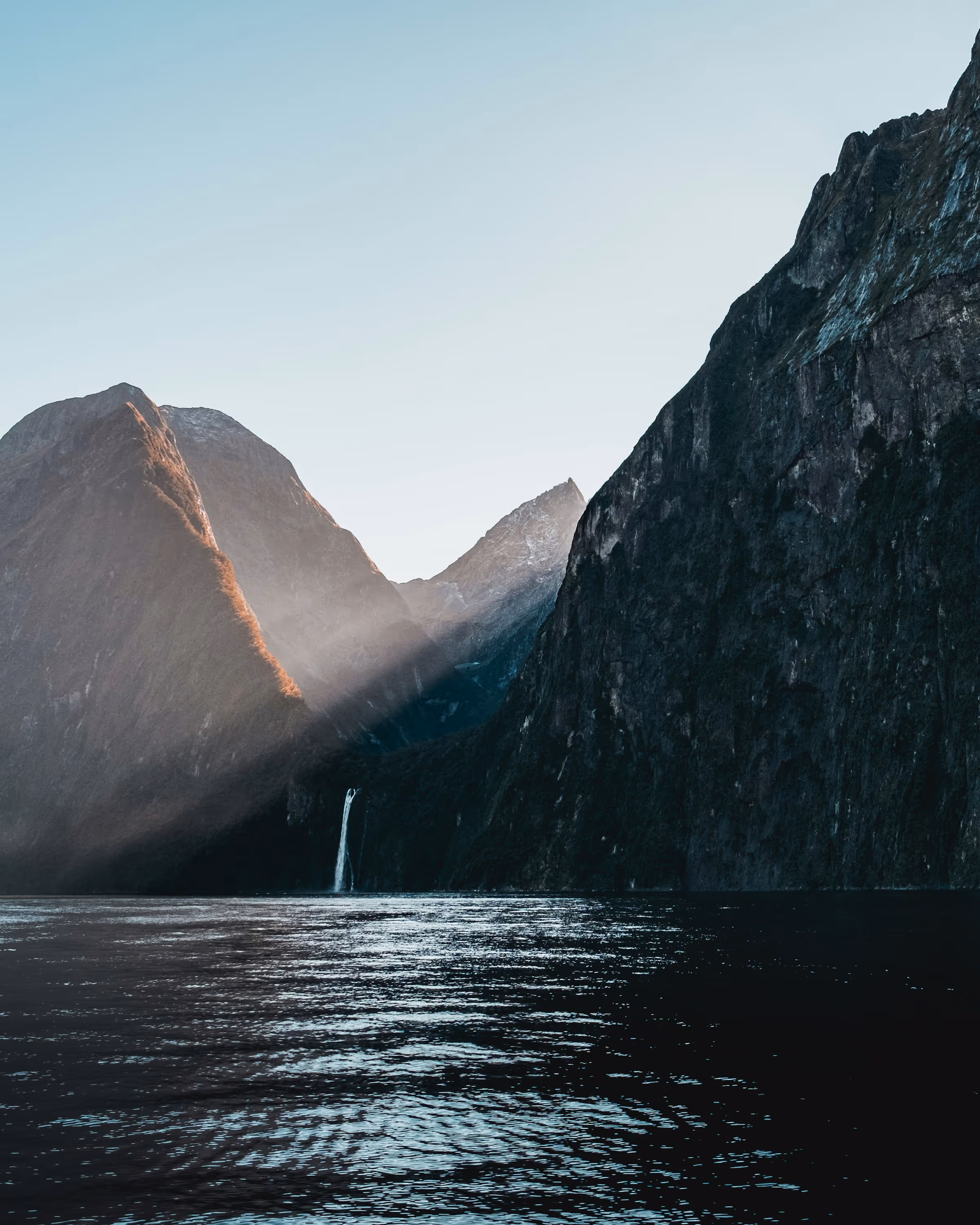Sunlight casting a beam over mountainous cliffs with a waterfall flowing into calm water.