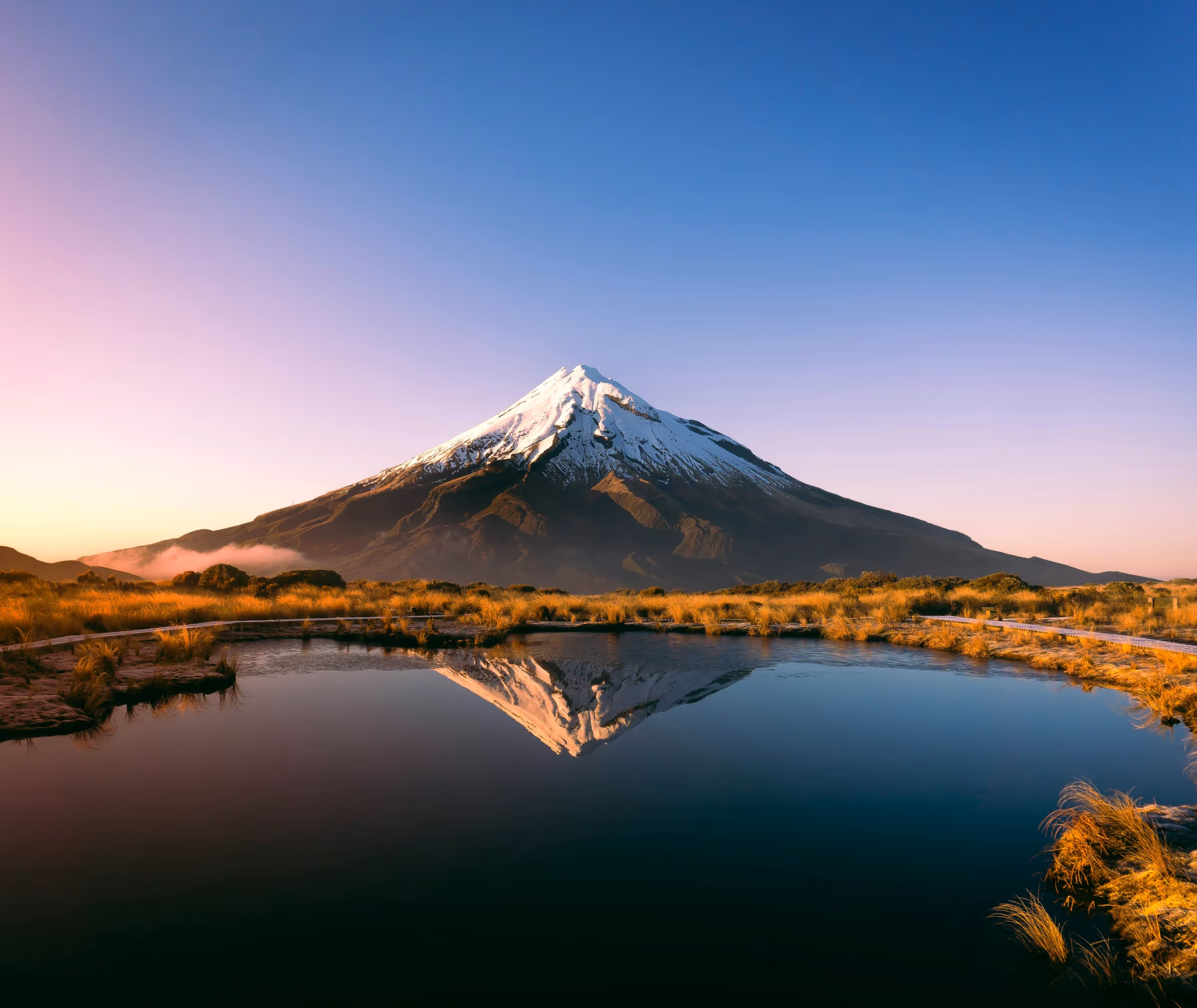 Snow-capped mountain reflected in a calm pond with golden grasses around at sunrise.