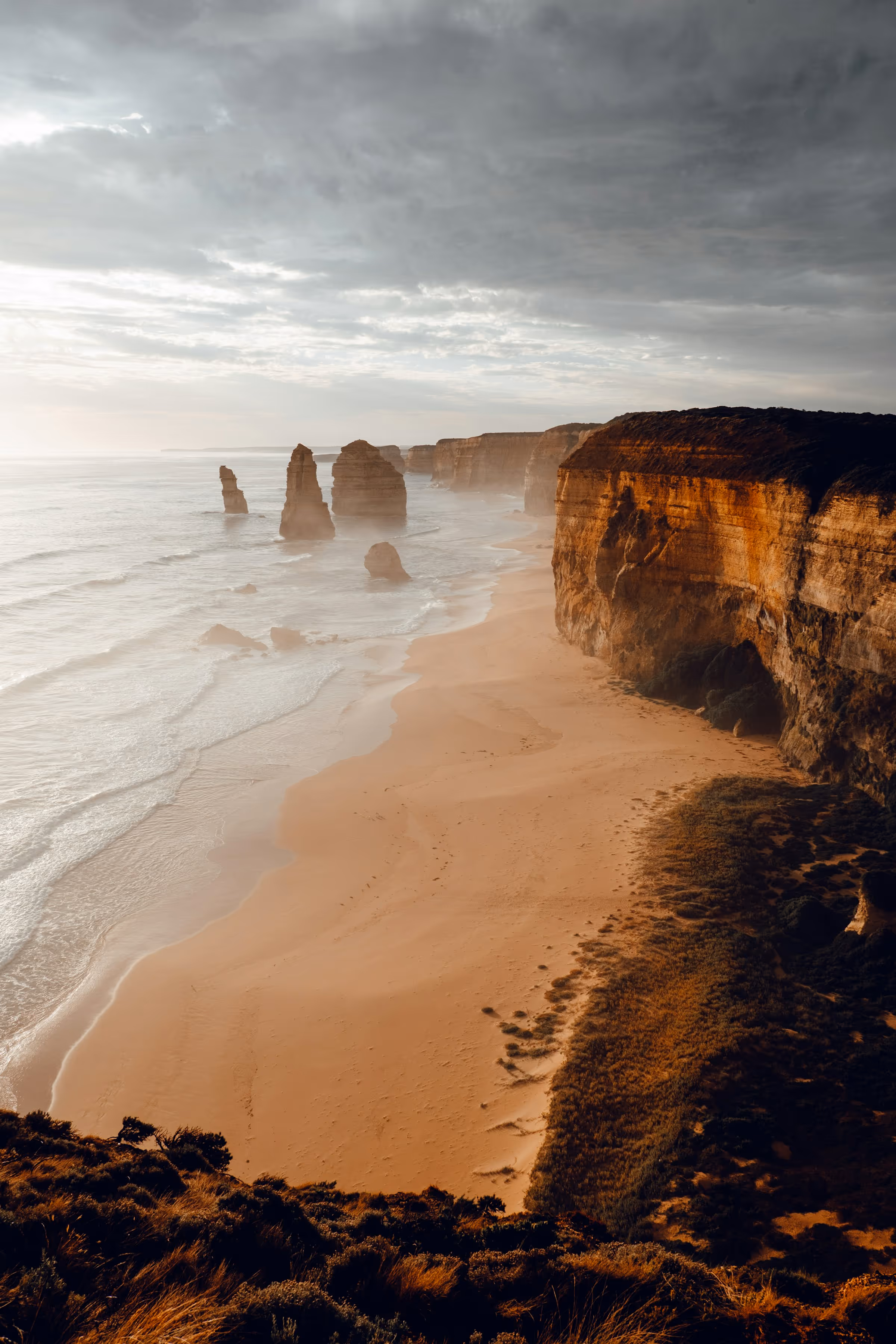 Coastal landscape with tall sea stacks rising from the ocean near sandstone cliffs and a sandy beach under a cloudy sky.