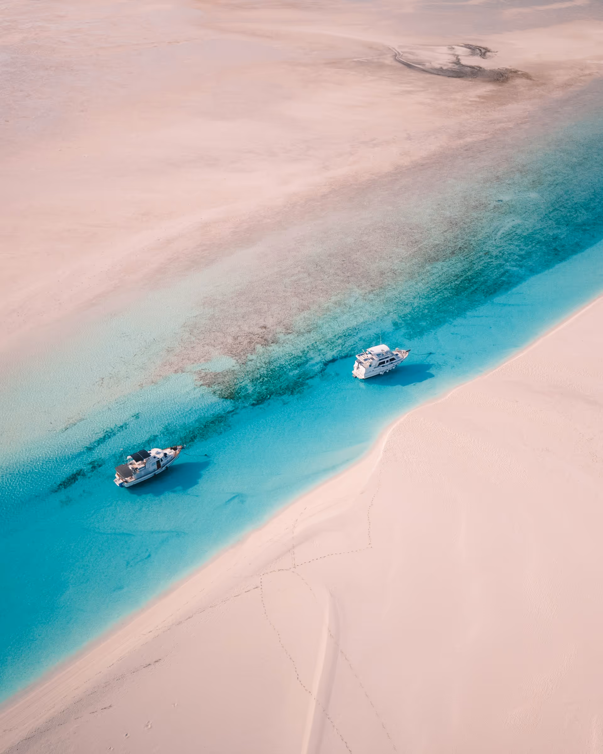 Aerial view of two boats anchored in clear turquoise water bordered by white sandy beach.