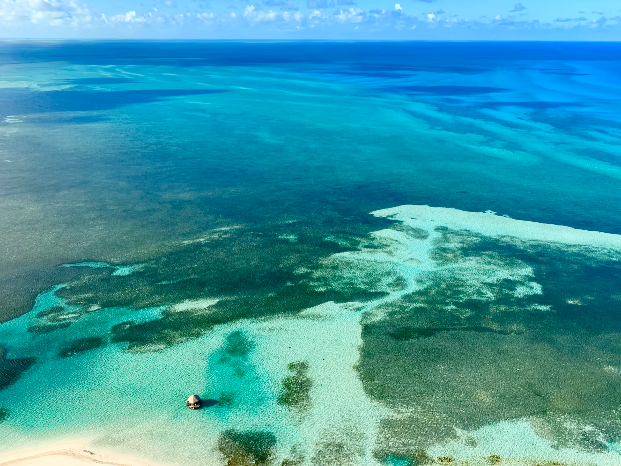 Aerial view of clear turquoise ocean water with sandbanks and a small hut on stilts near the shore.