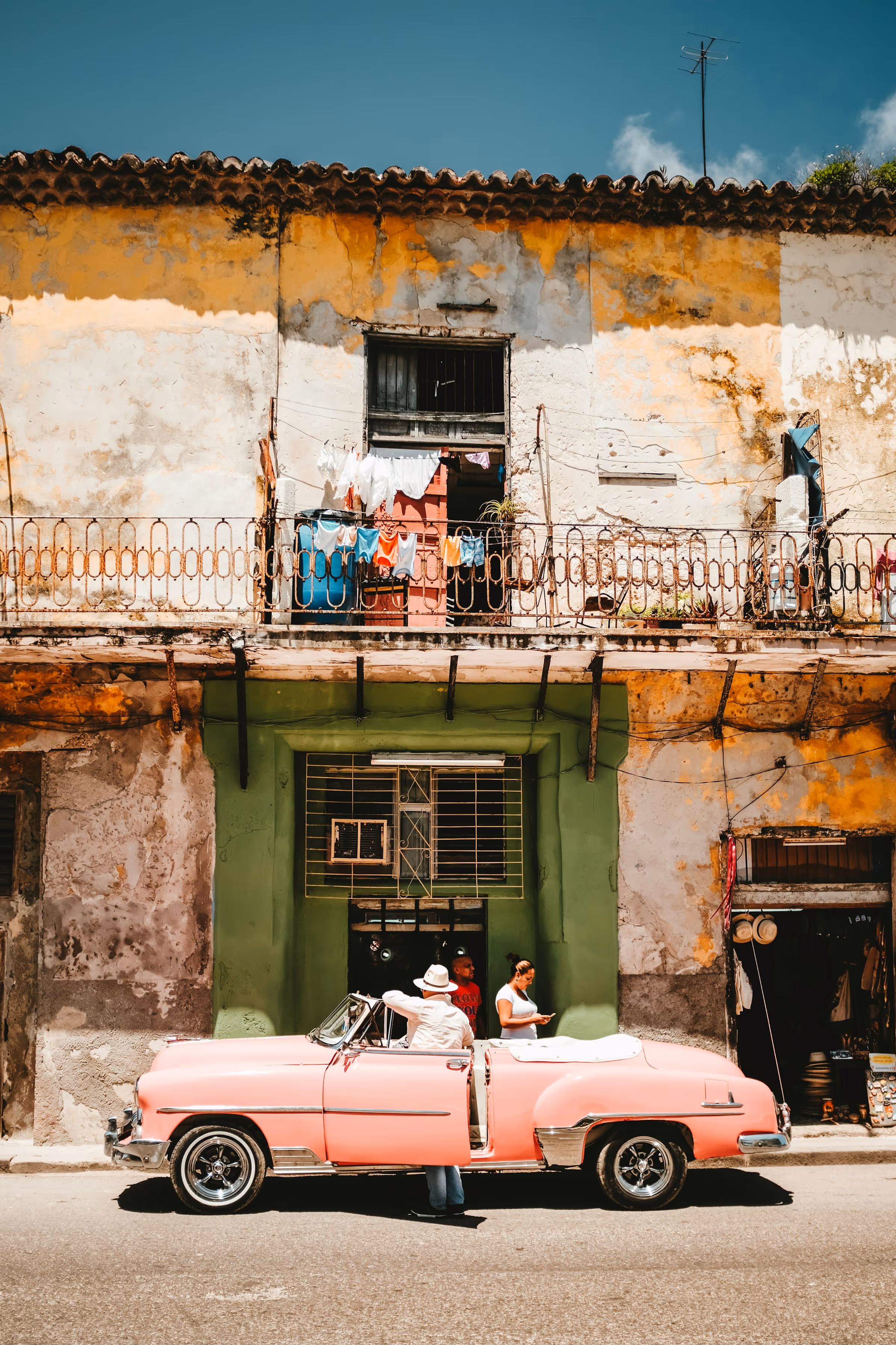 Vintage pink convertible car parked on a sunny street in front of a weathered yellow and green building with people around it.