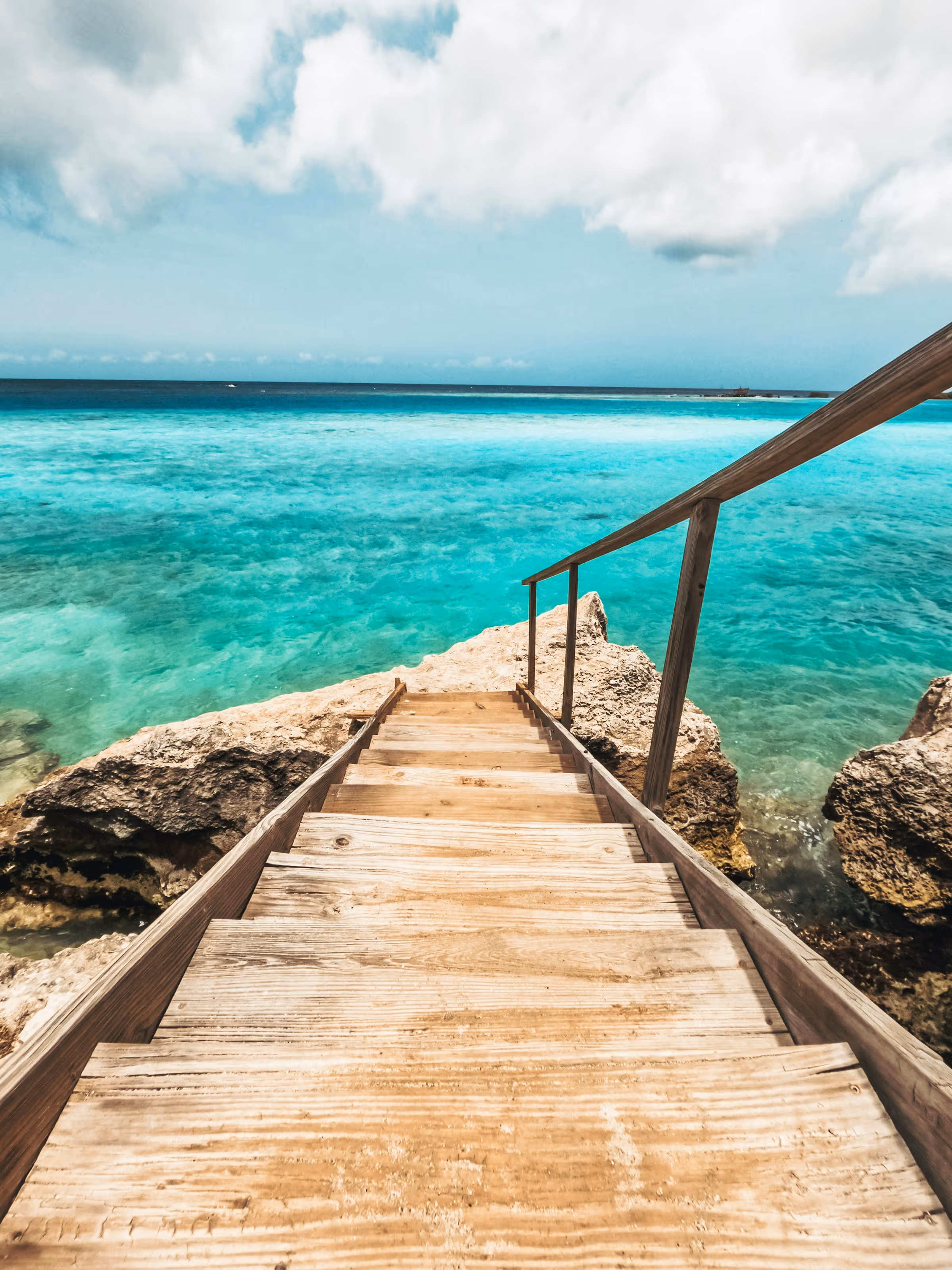 Wooden stairs with railings leading down to clear turquoise ocean water and rocky shoreline under a partly cloudy sky.