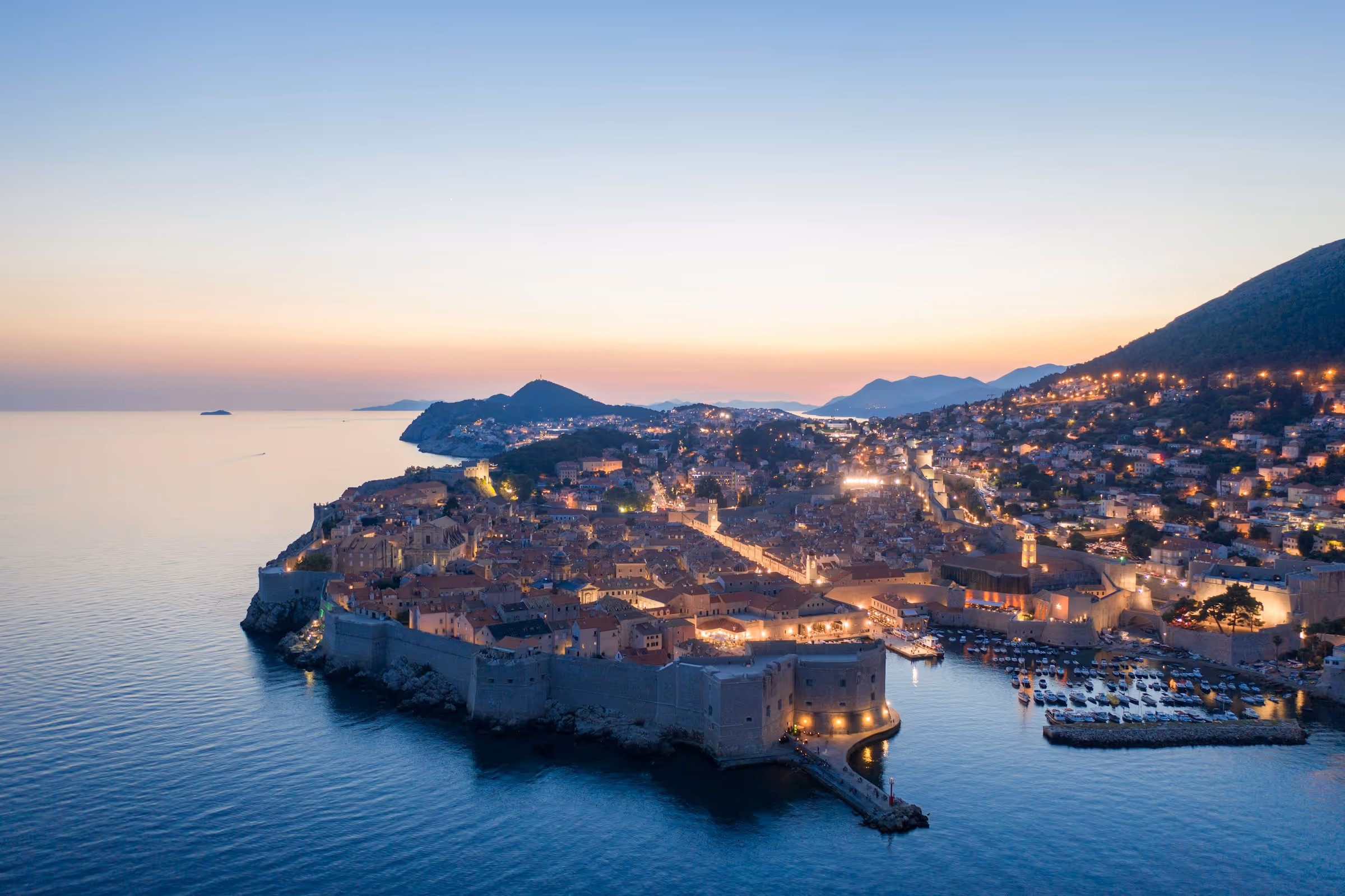 Aerial view of a coastal city at dusk with illuminated historic buildings, city walls, and a harbor filled with boats.