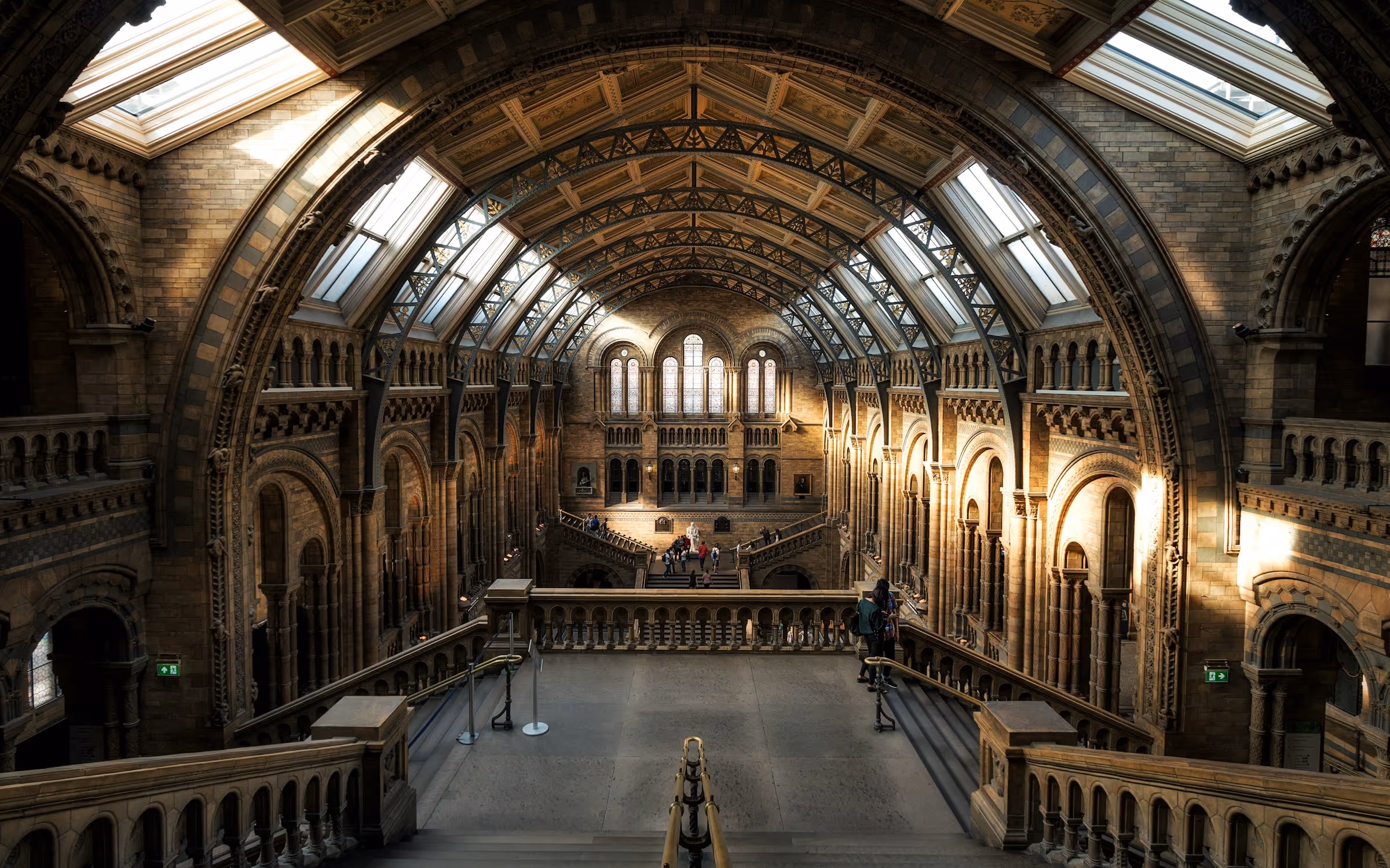 Interior of a grand historic building with vaulted ceilings, arched windows, and a wide staircase overlooking visitors below.