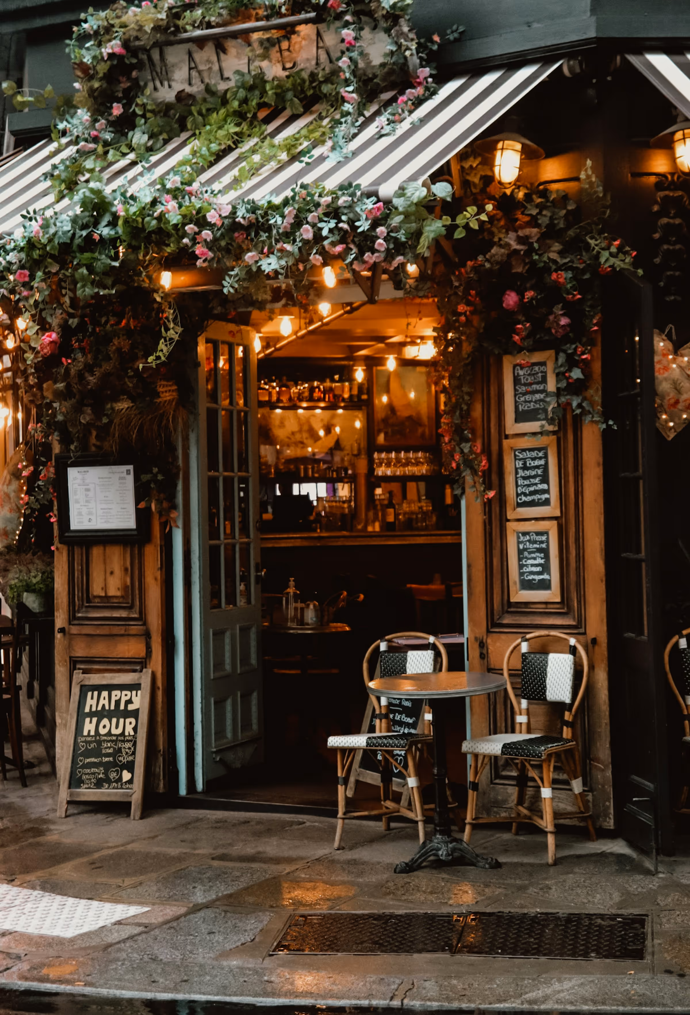 Cozy café entrance decorated with hanging plants and flowers, featuring a small table and two chairs on a wet stone pavement.