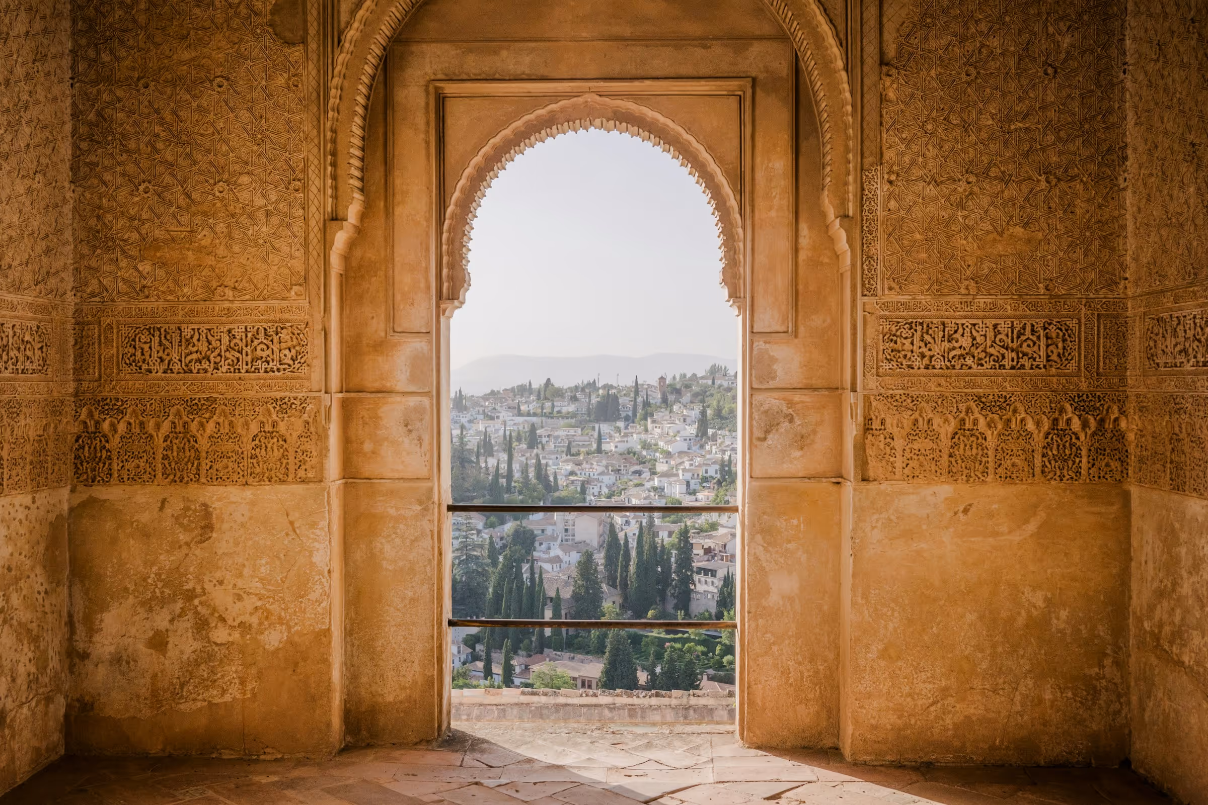 View of a city with white buildings and cypress trees framed by an ornate arched stone window.