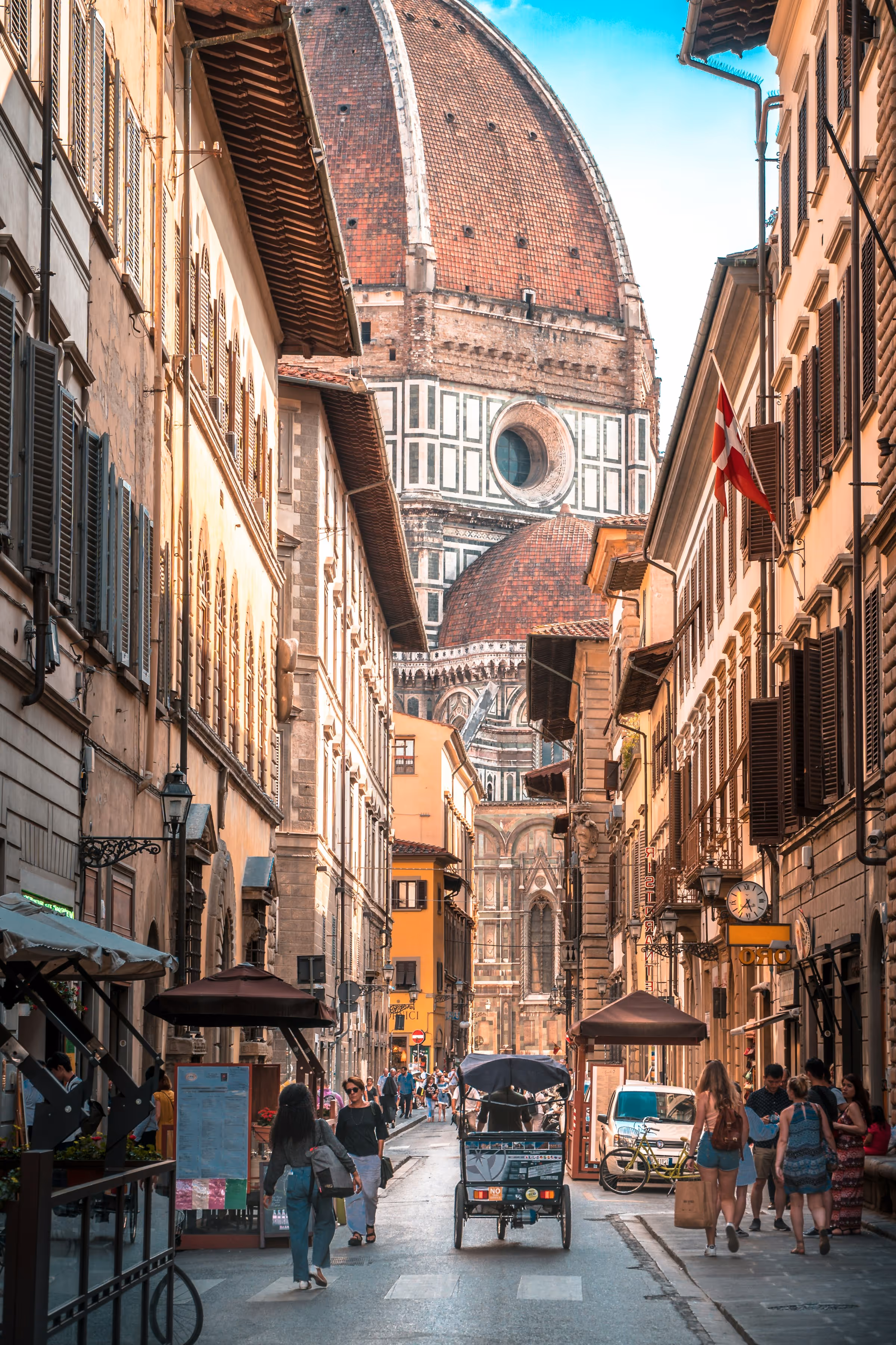 Busy street in Florence, Italy, with people walking and riding a rickshaw, and the dome of Florence Cathedral visible in the background.