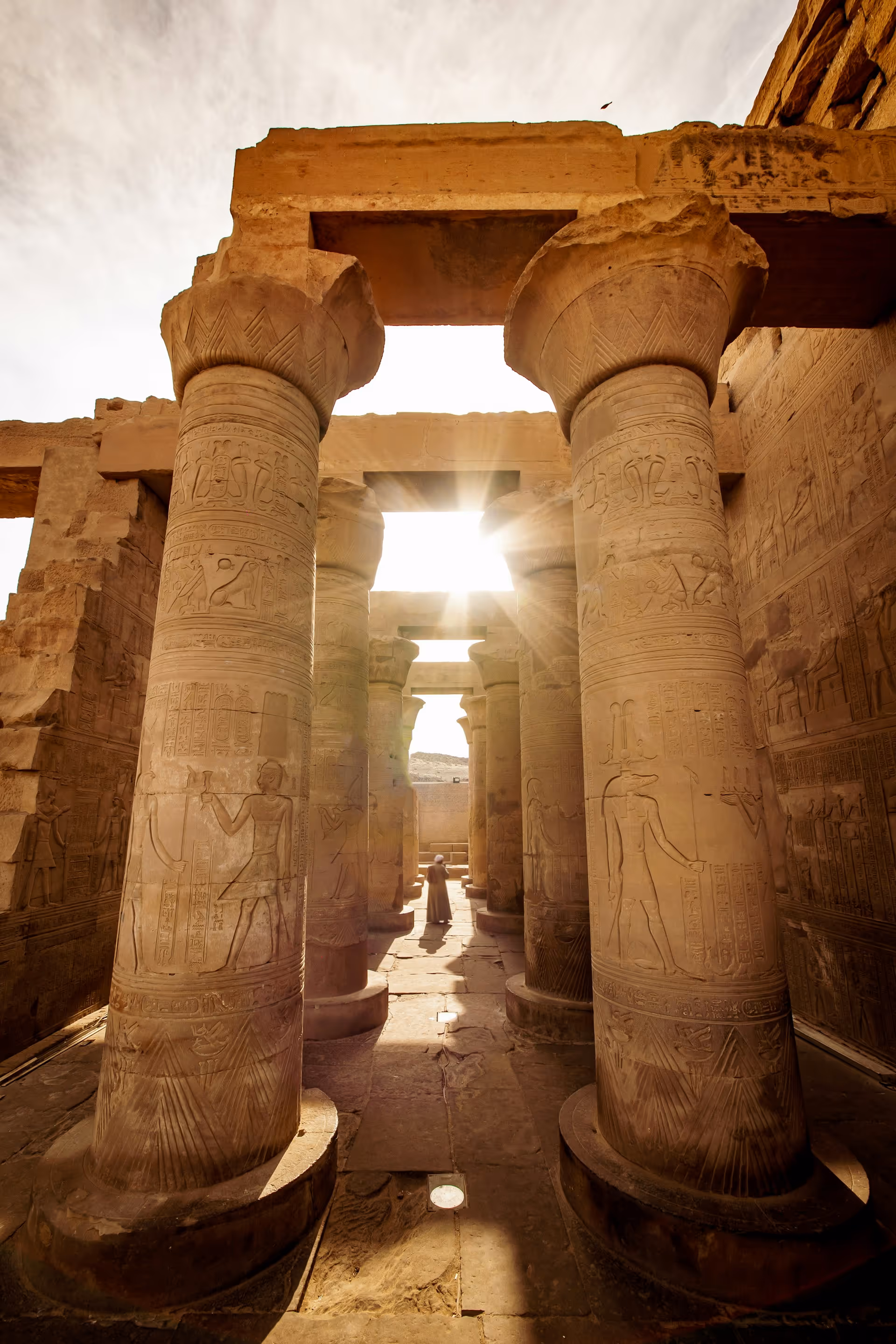 Sunlight shining through ancient Egyptian temple pillars carved with hieroglyphics and figures, with a person walking in the distance.