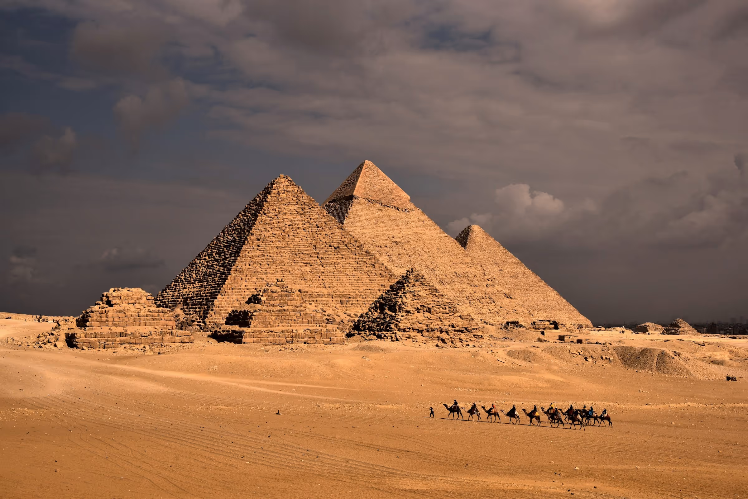 The Pyramids of Giza in Egypt with a caravan of camels and riders crossing the sandy desert in the foreground under a cloudy sky.
