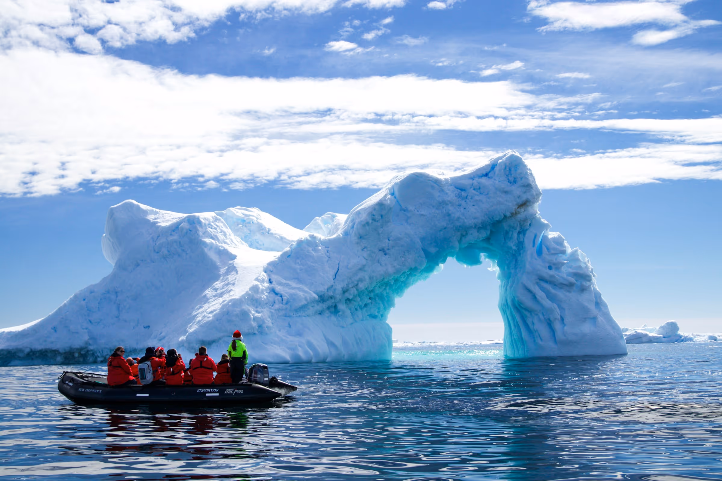 Group of people in red jackets on a small black inflatable boat near a large ice arch floating in calm ocean water under a partly cloudy sky.