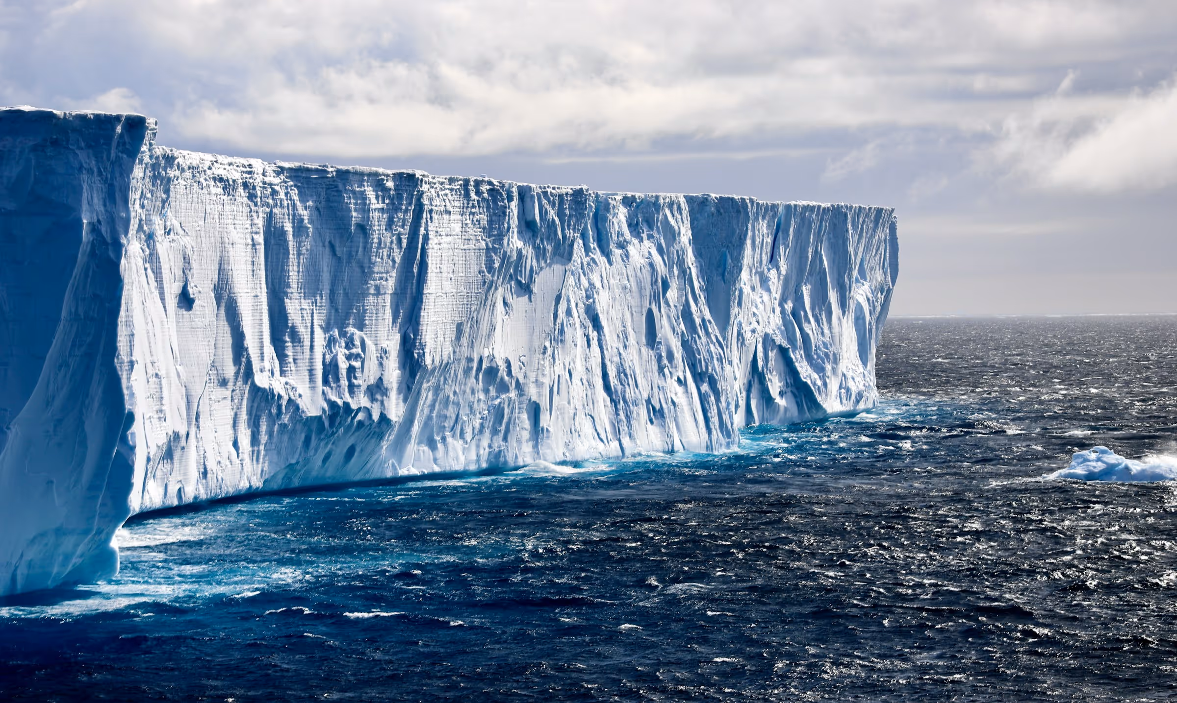 Large ice shelf towering over dark blue ocean waters under a cloudy sky.