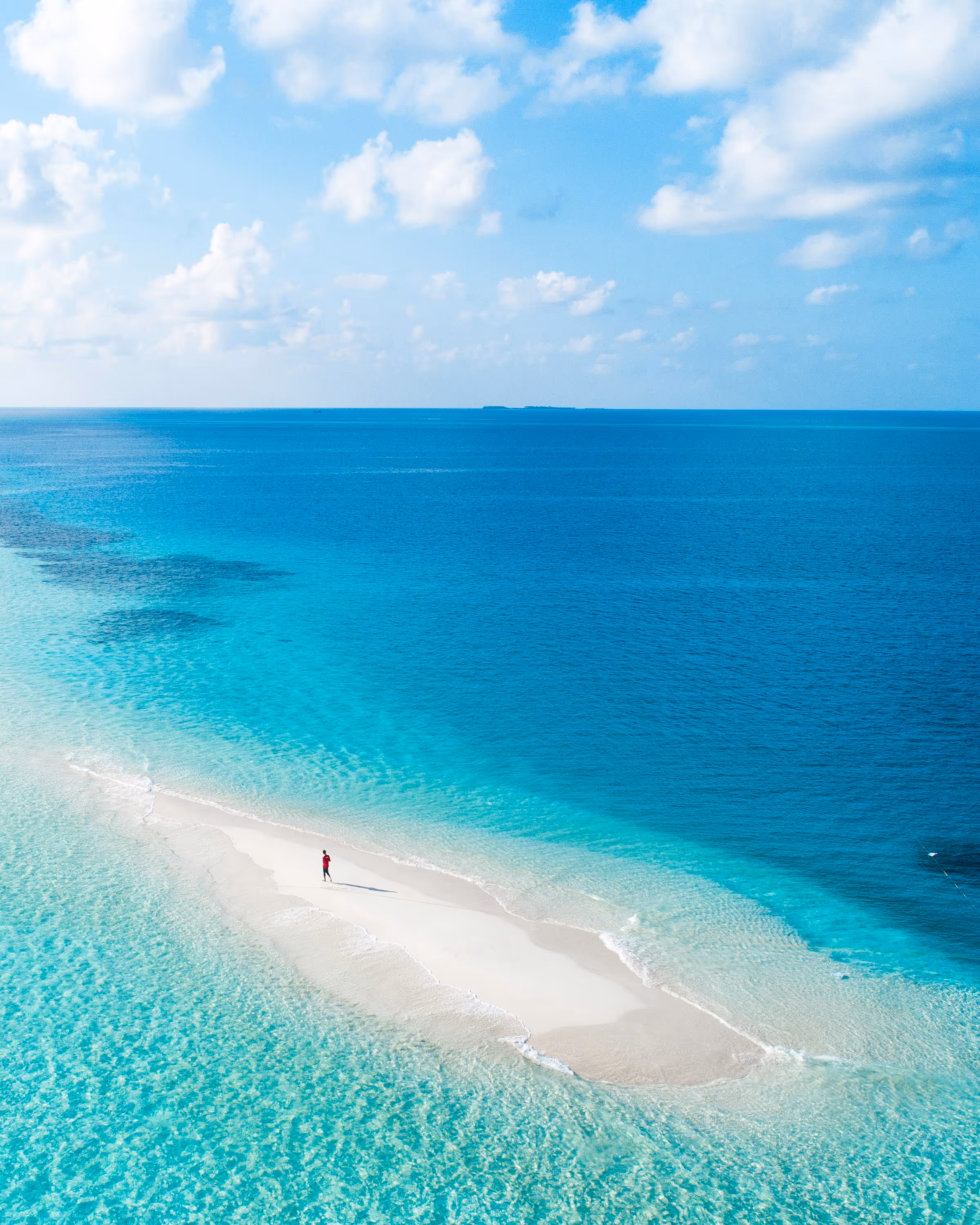 Aerial view of a small sandy island with a single person in a red shirt standing, surrounded by clear turquoise and deep blue ocean under a partly cloudy sky.