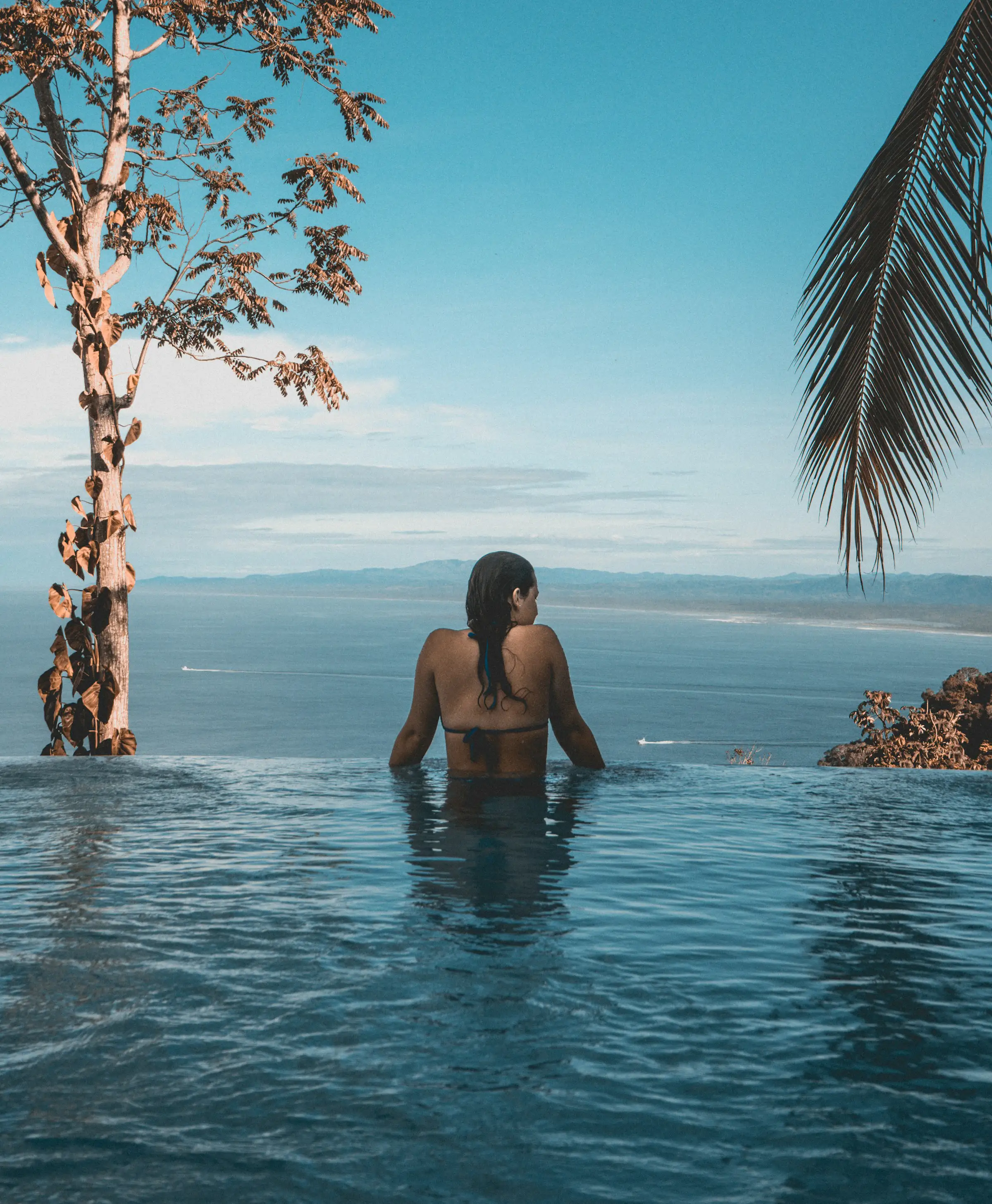 Woman in a bikini relaxing in an infinity pool overlooking a vast ocean and distant mountains under a clear blue sky.