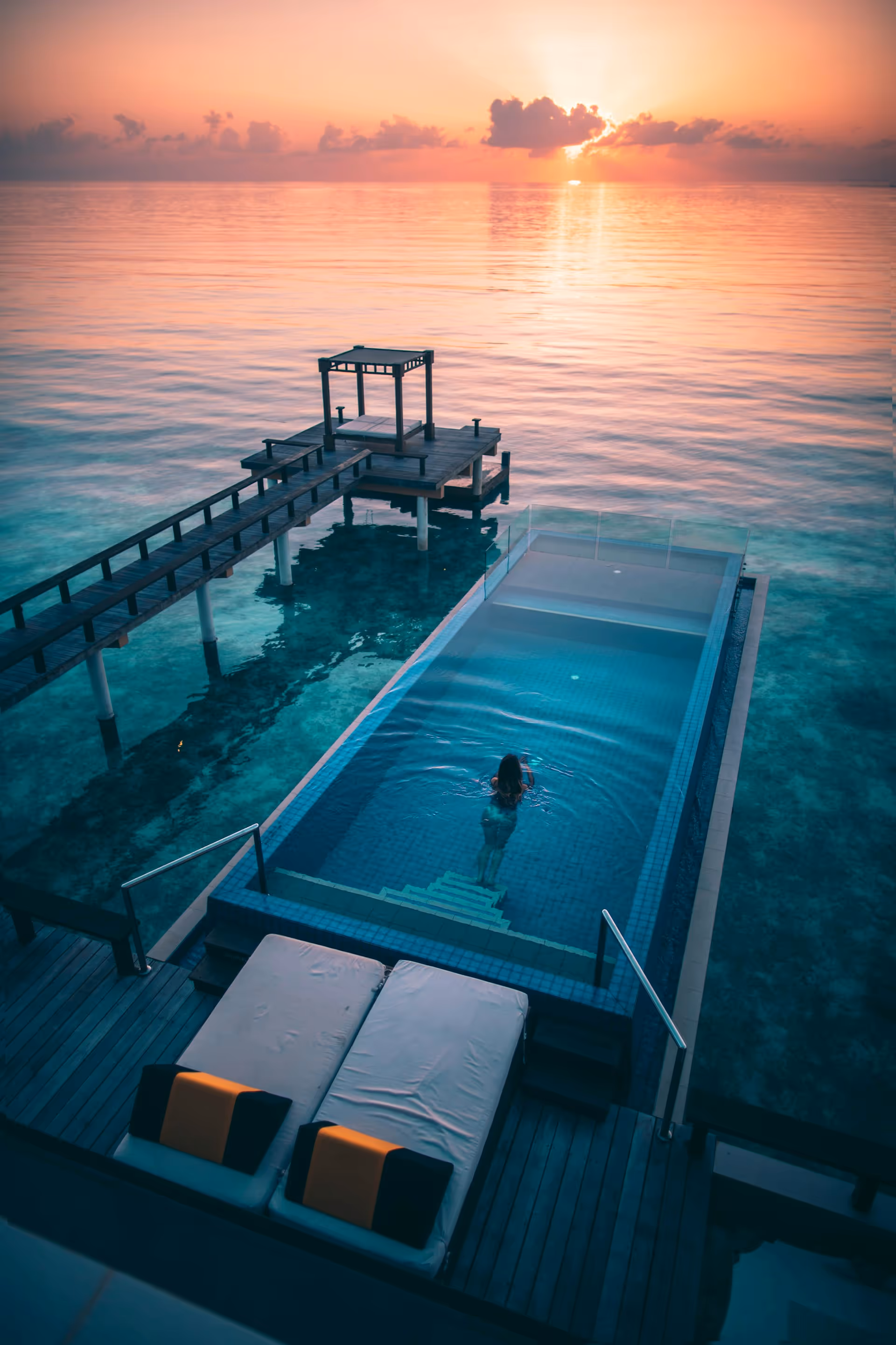 Person swimming in an infinity pool overlooking a wooden pier and calm ocean at sunset.