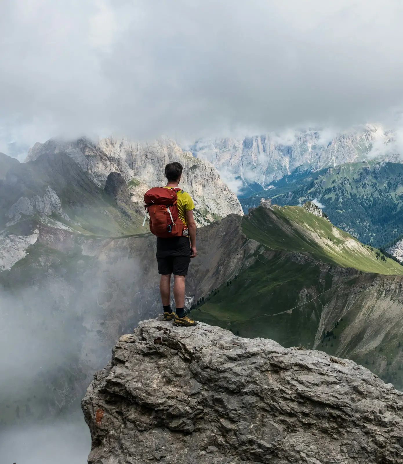 Hiker with a red backpack standing on a rocky cliff overlooking misty mountains and green valleys.