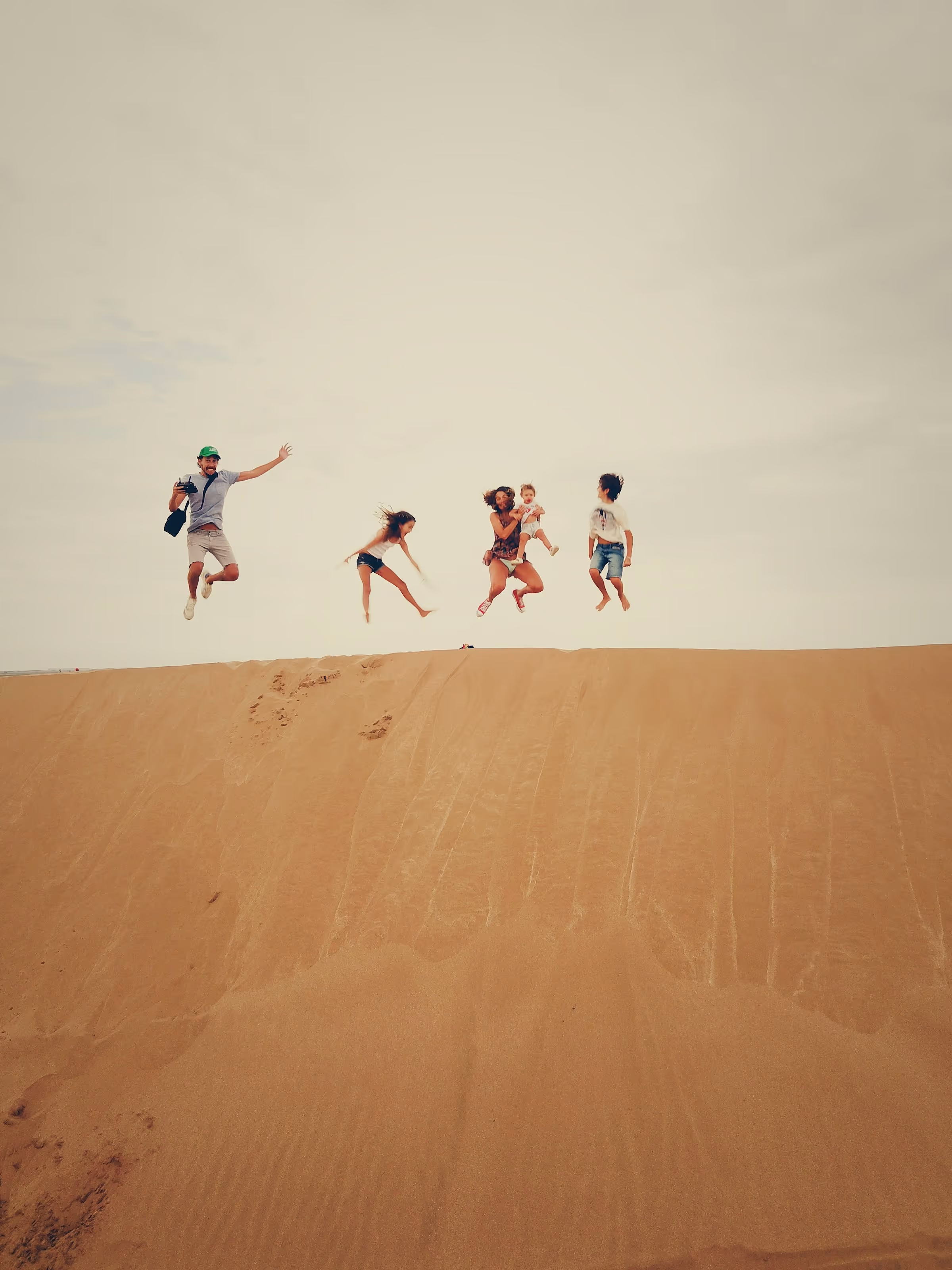 A man and four children jumping mid-air on sand dunes under a cloudy sky.