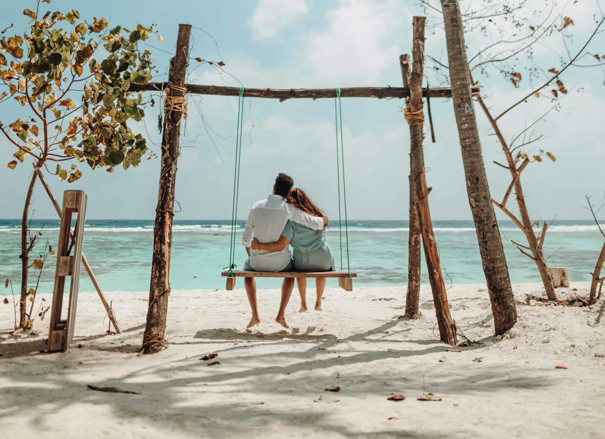 Couple sitting closely on a wooden swing by the beach, overlooking the ocean with a partly cloudy sky.