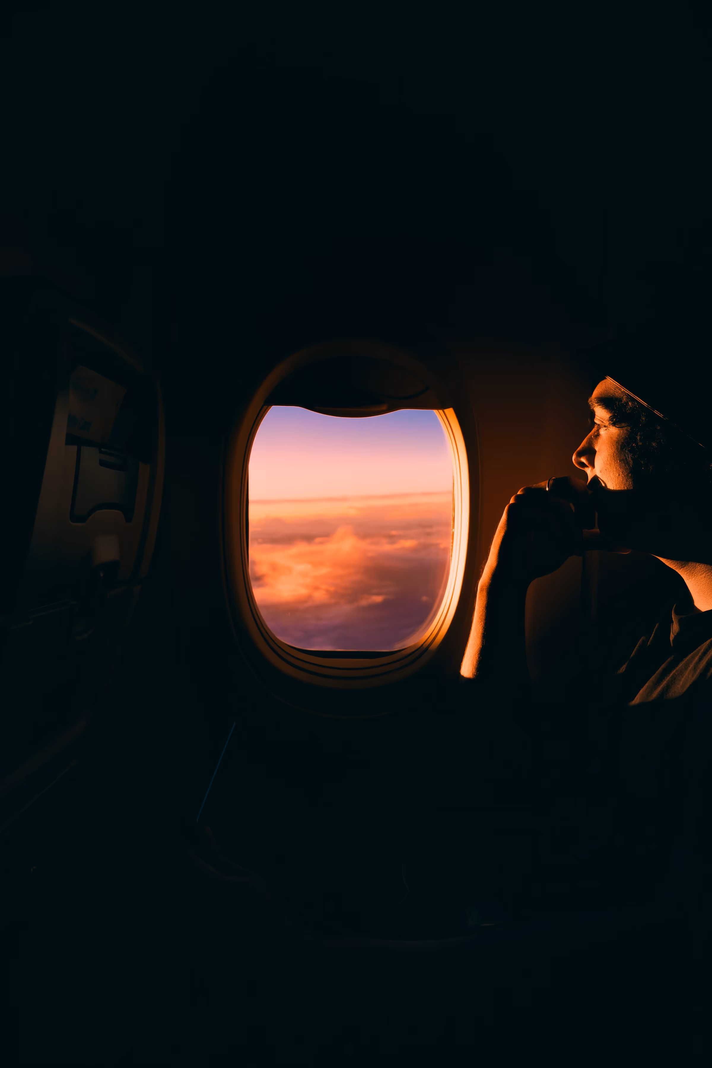 Person looking thoughtfully out of an airplane window at a sunset with clouds.