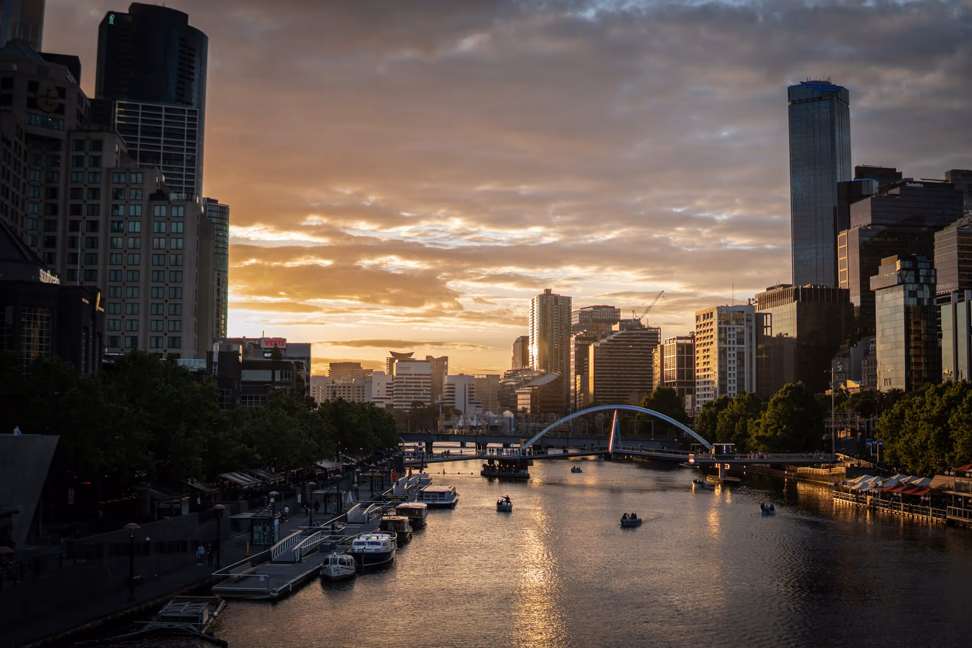 Sunset over a river in a city with modern high-rise buildings, boats on the water, and a pedestrian bridge.