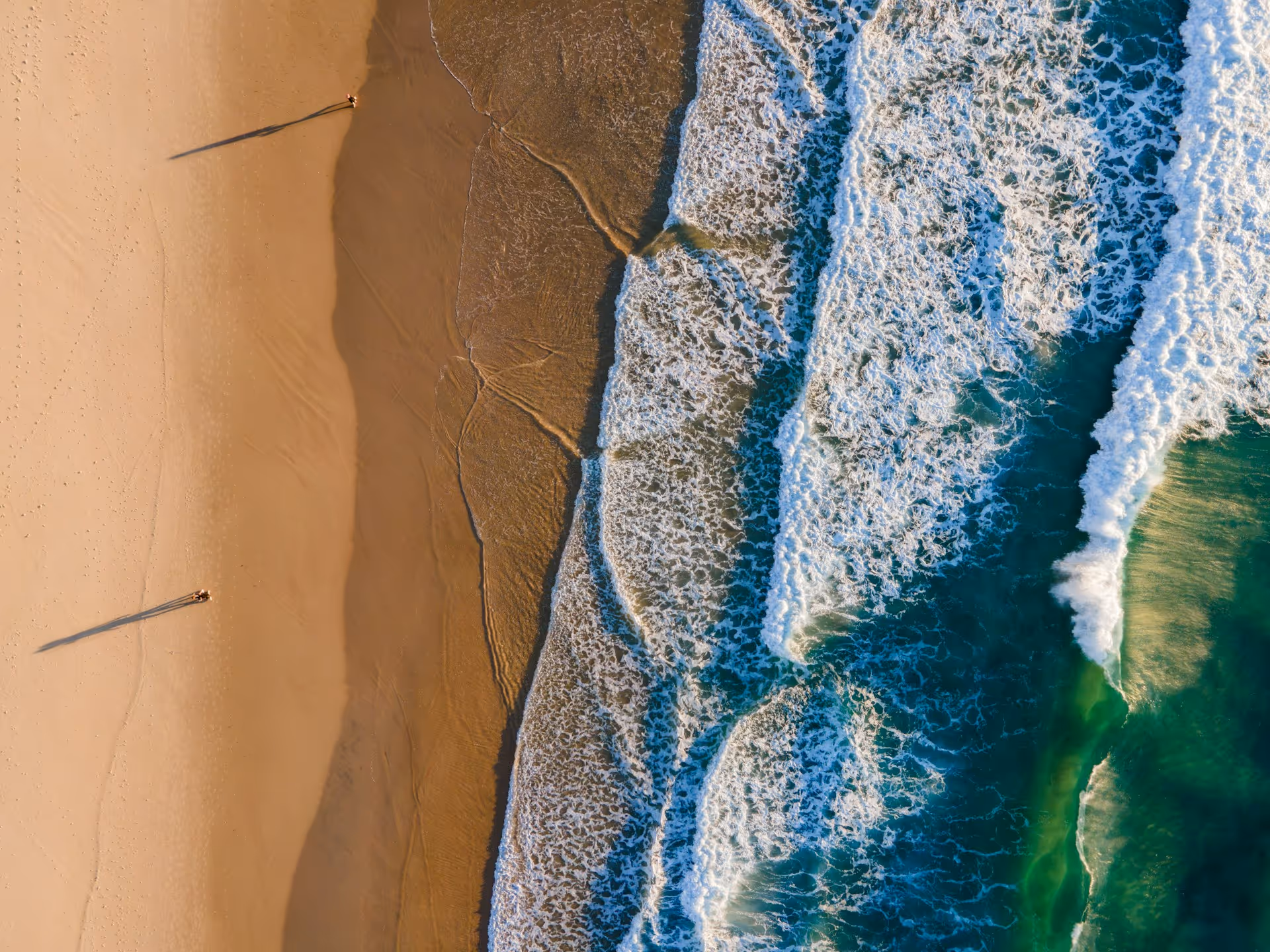 Aerial view of two people standing apart on a sandy beach with long shadows, near foamy ocean waves.