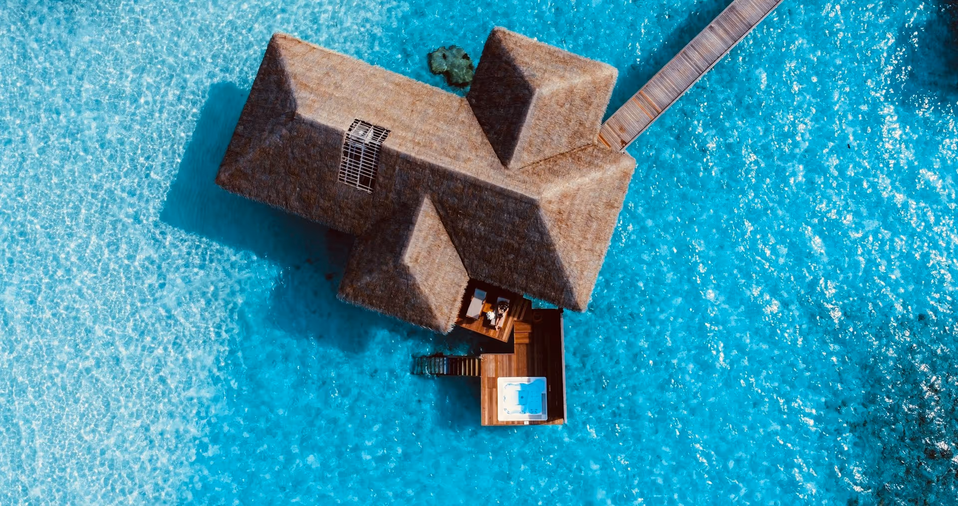 Aerial view of a thatched-roof overwater bungalow with a wooden deck and jacuzzi in clear turquoise ocean water.