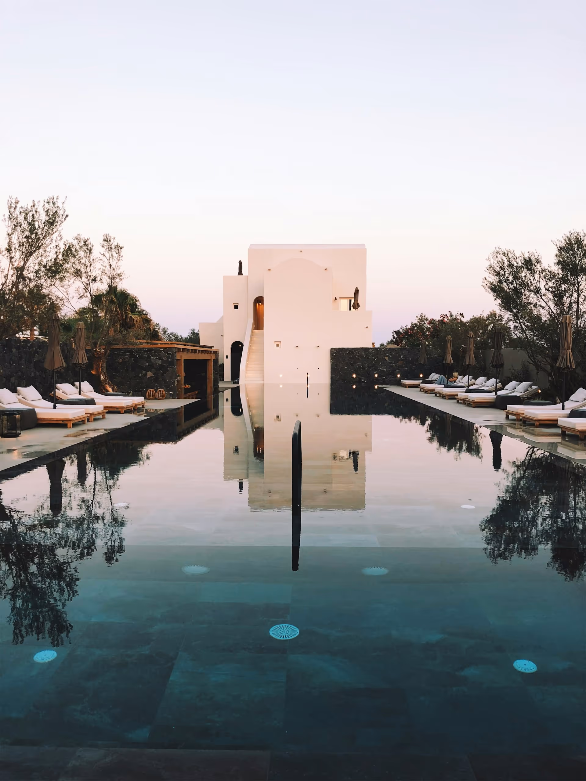 Modern white building reflected in a calm swimming pool surrounded by lounge chairs and trees at dusk.
