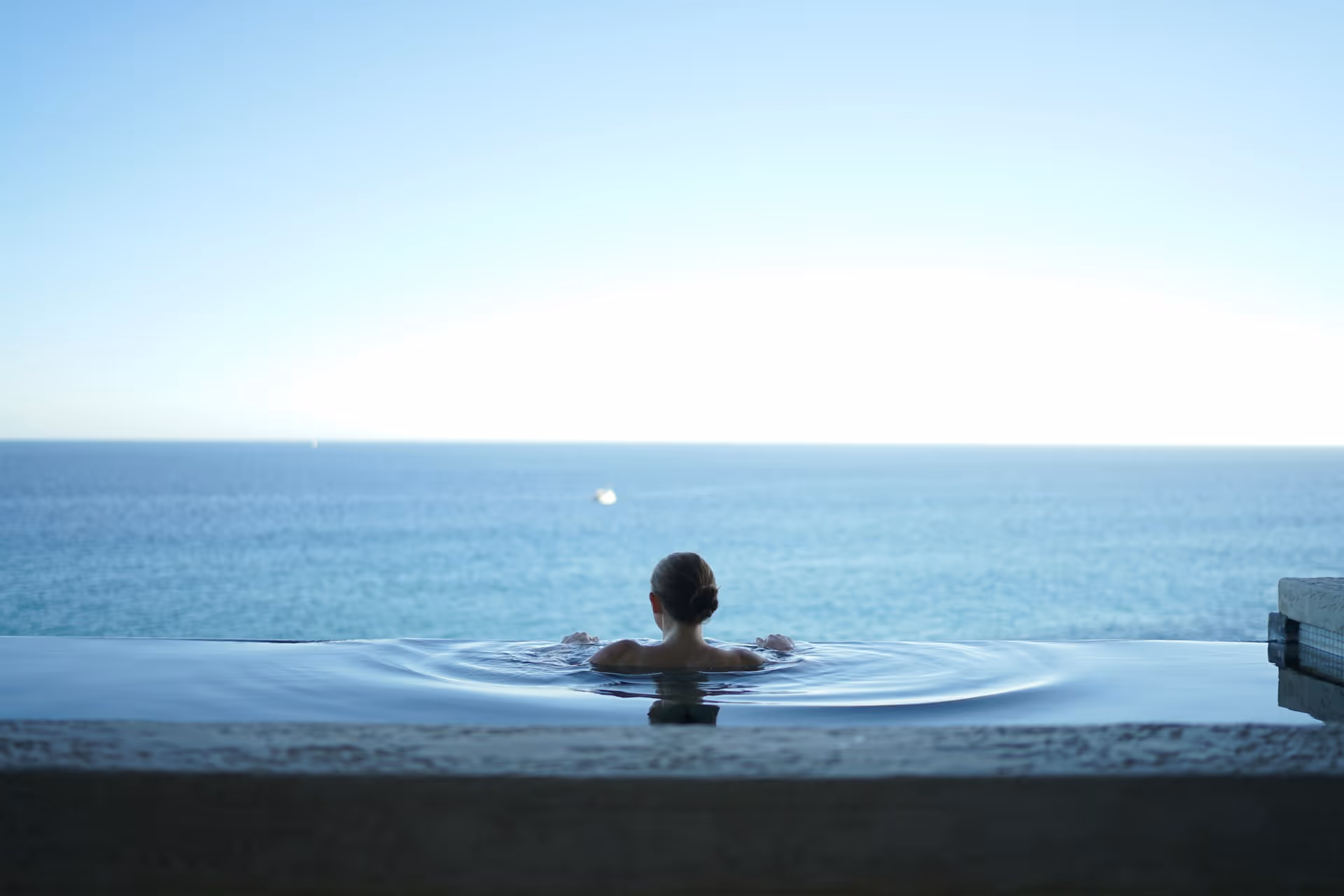 Person relaxing in an infinity pool overlooking a calm ocean under a clear sky.